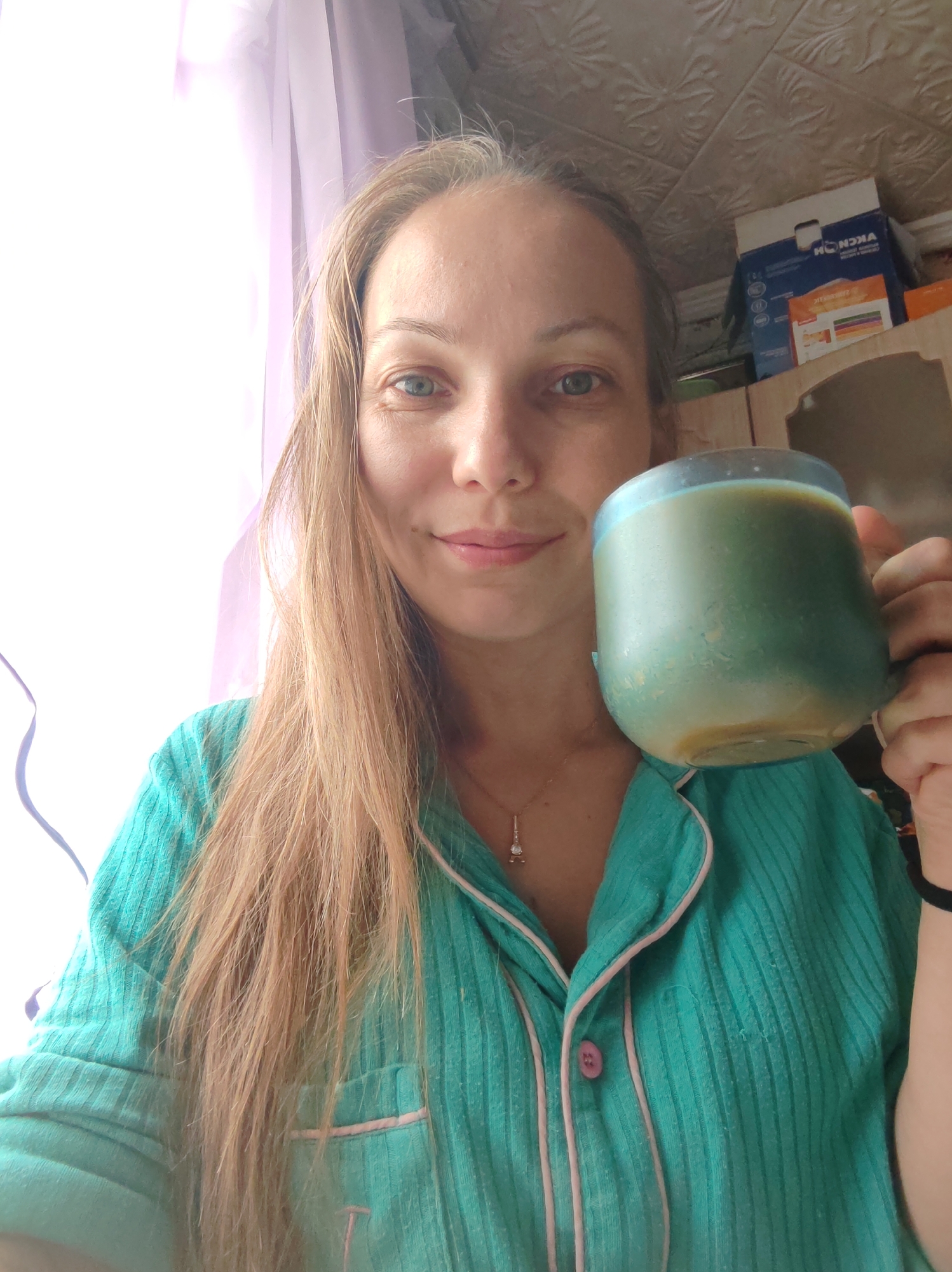 A woman with long blonde hair, wearing a teal pajama top with pink trim and buttons, holds a blue and green mug filled with a beverage in her right hand. She is looking at the camera and smiling slightly, with her head tilted. The background shows a window with sheer purple curtains on the left, and shelves with boxes on the right. The ceiling, visible at the top of the image, appears to be a textured wallpaper. A small, diamond-like pendant hangs from a necklace on the woman's neck. The lighting suggests it is daytime, with natural light coming from the window. The overall impression is a casual, domestic scene.