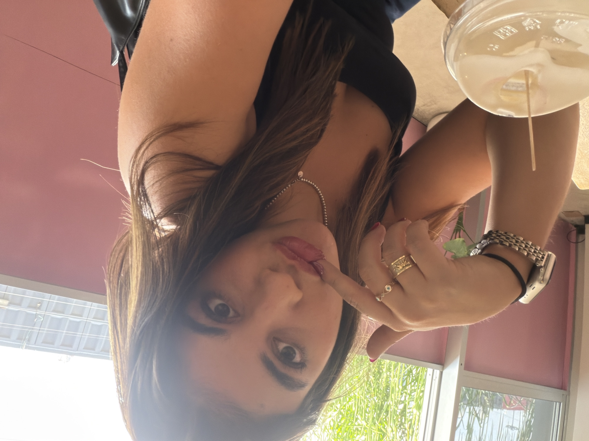A woman poses with her finger touching her lips. She is wearing a black top and a necklace. She has multiple rings and a watch on her wrist. A glass with a drink and straw is visible. The location appears to be indoors near a window with a view of green vegetation. The image is potentially taken in Puebla, Mexico.