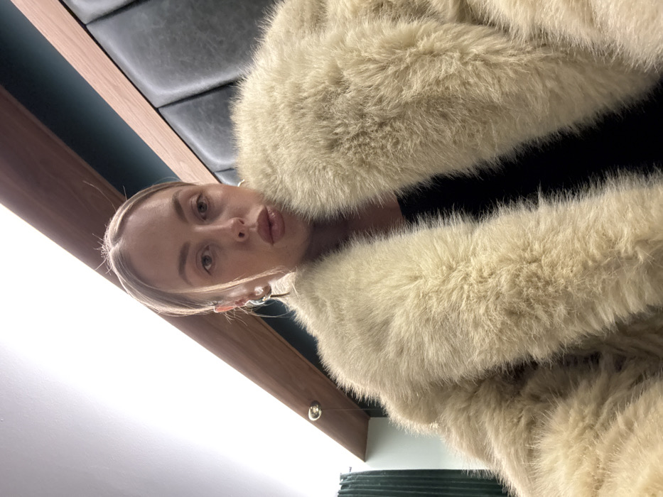 A woman poses for a low-angle selfie, her face close to the camera with slightly puckered lips. She is surrounded by a fluffy, cream-colored fur coat. Behind her, a dark tufted wall meets a wood-paneled ceiling with recessed lighting. The overall perspective is somewhat disorienting due to the tilted angle.