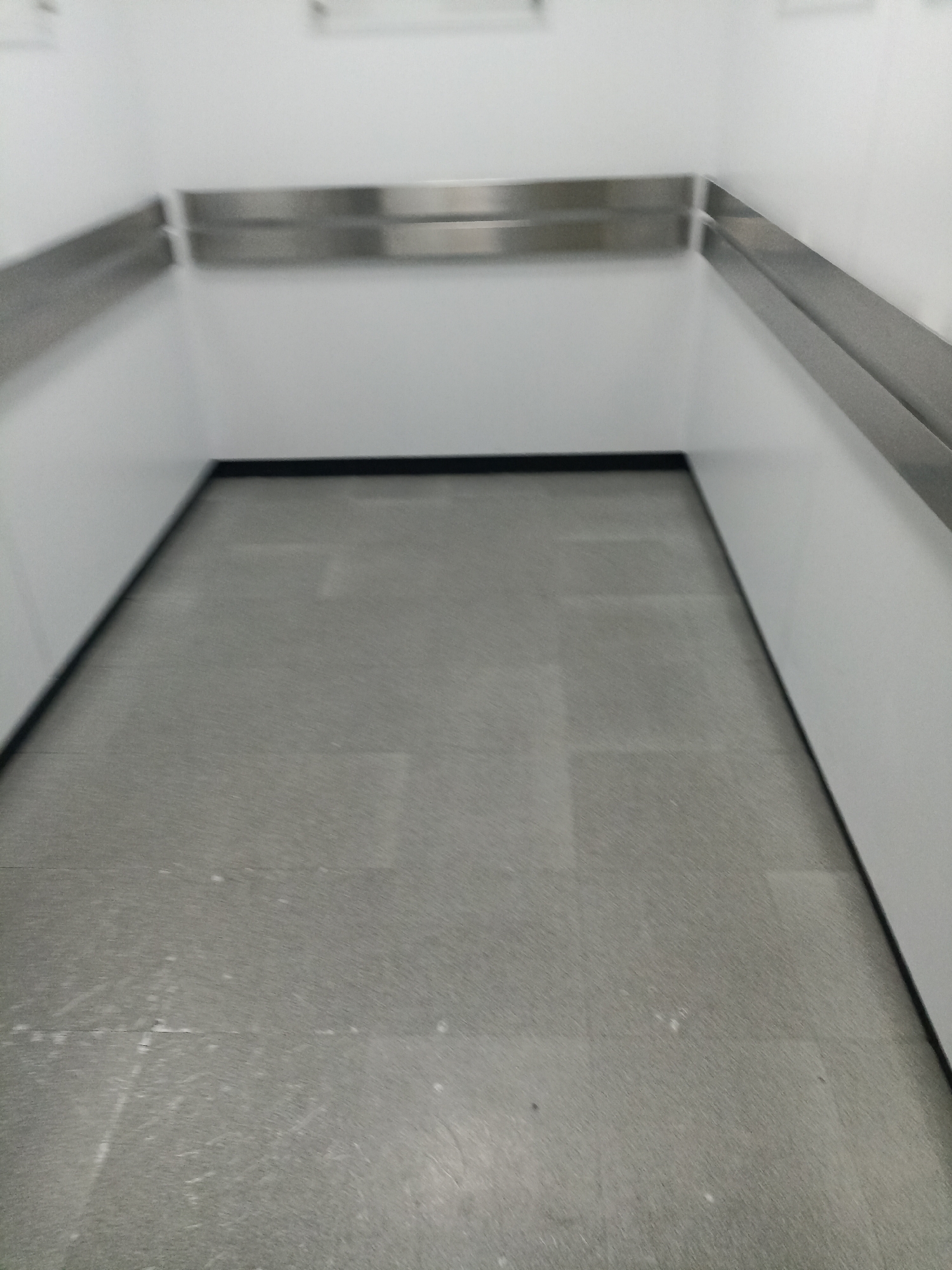 The image depicts an empty, indoor space with a grey tiled floor and white walls. A continuous, horizontal stainless steel bar runs along the top of the walls at approximately waist height. The space appears to be a walkway or a narrow room. There is no indication of the specific location within Surabaya, Indonesia, or any people or objects other than the architectural elements.