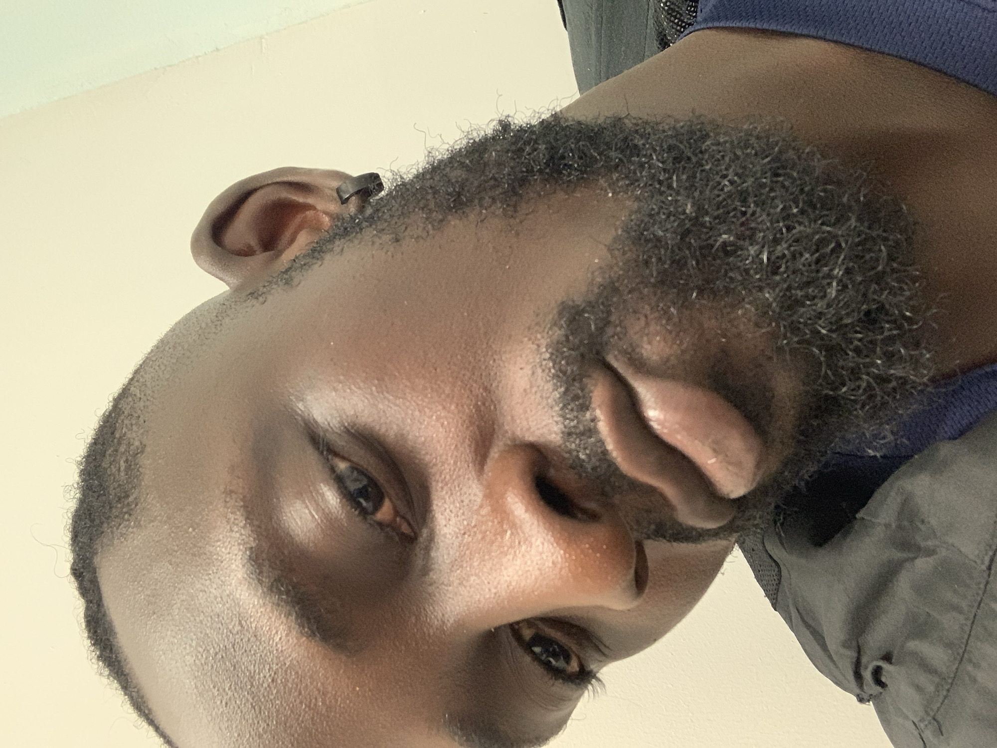 Close-up, angled view of a dark-skinned male's face. His head is positioned so that the top of his head is near the left side of the frame. The man has a short, curly beard. His eyes are dark and focused toward the camera. His nose is centered, with his lips pursed. A portion of his ear is visible on the left side of the frame. He appears to be wearing a navy-blue top under a gray jacket. The background is a plain, pale color. No indication of the location being Lekki, Nigeria, is visible in the image.