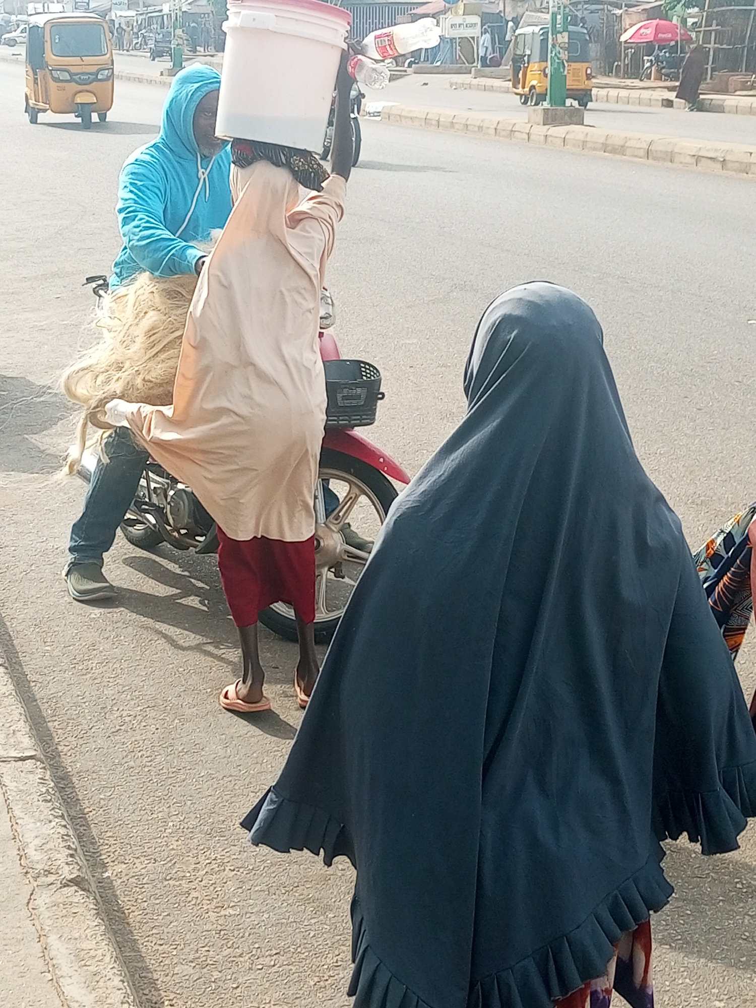 A woman in a dark blue hijab walks away from the camera on a paved road in Tempure, Nigeria. She is wearing a ruffled garment underneath the hijab, and her feet are adorned with orange flip-flops. The road is lined with a concrete curb. In the background, a man on a motorcycle wears a bright blue hoodie and jeans. He is transporting a large bundle of straw or hair, which spills out from behind him. On his head, he balances a large white bucket. To the left of the motorcycle, a yellow auto-rickshaw drives away from the camera. In the distance, several buildings and a red umbrella are visible, suggesting a market or roadside businesses. The sun appears to be bright, casting shadows, indicating it is likely daytime.