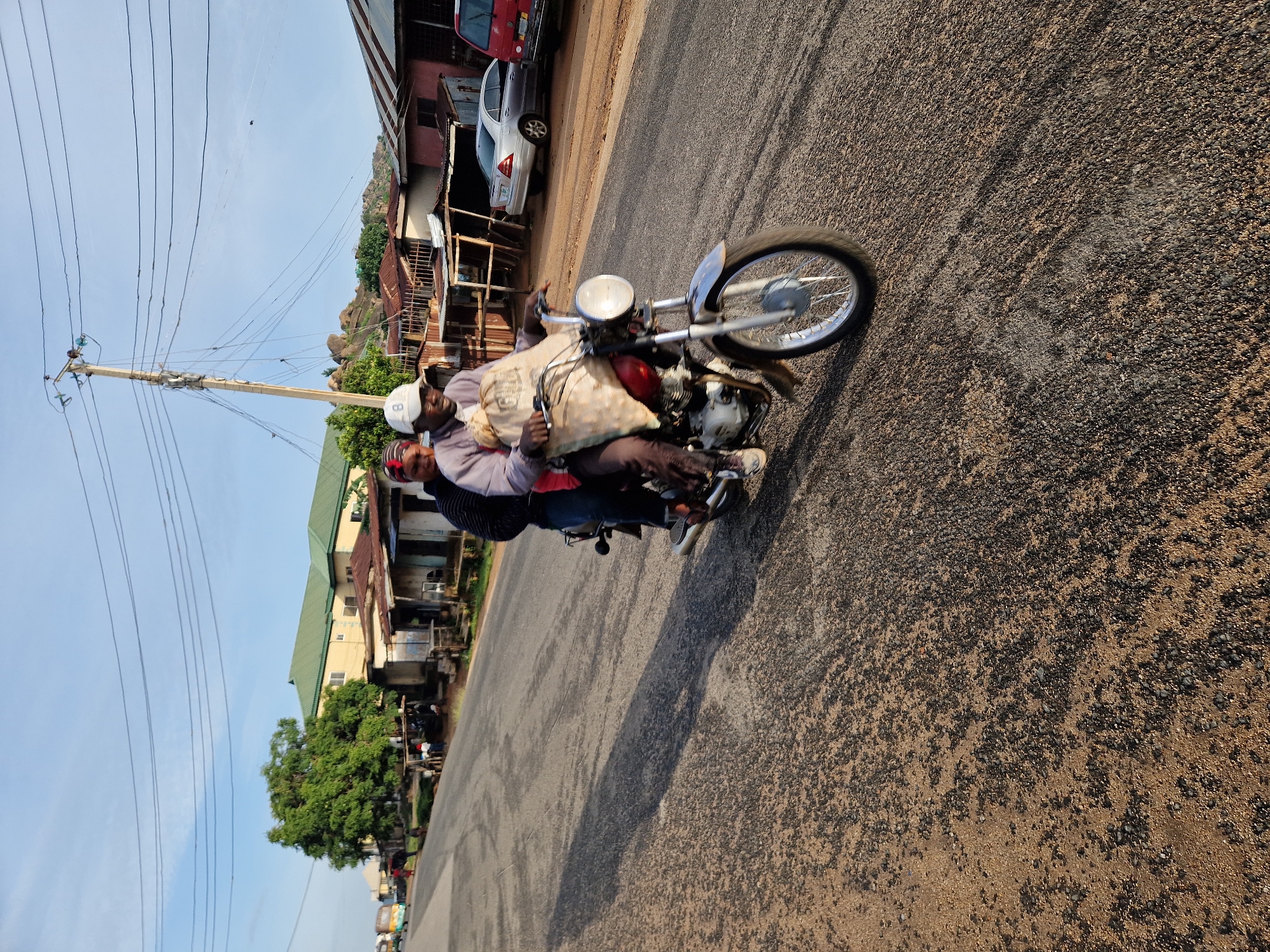 Two individuals are riding on a motorcycle on a paved road in Rukuba Road, Nigeria. The person in the front is operating the motorcycle and appears to be wearing a light-colored long-sleeved shirt, dark pants, and a white baseball cap. They are carrying a large, beige sack over their lap and shoulders. The person behind them is wearing a dark-colored long-sleeved shirt and a red head covering. Both individuals are facing forward, suggesting they are traveling along the road. The motorcycle is a standard model with a visible engine, front wheel, headlight, and rear wheel. The road surface is asphalt with visible gravel and texture. To the right of the road, there are buildings with corrugated metal roofs, some appearing to be small shops or dwellings. A silver sedan and a red vehicle are parked on the right side of the road. In the background, there are trees, hills, and a utility pole with multiple wires. The sky is clear and blue, indicating daylight. The overall scene depicts everyday transportation and street life in a Nigerian city.