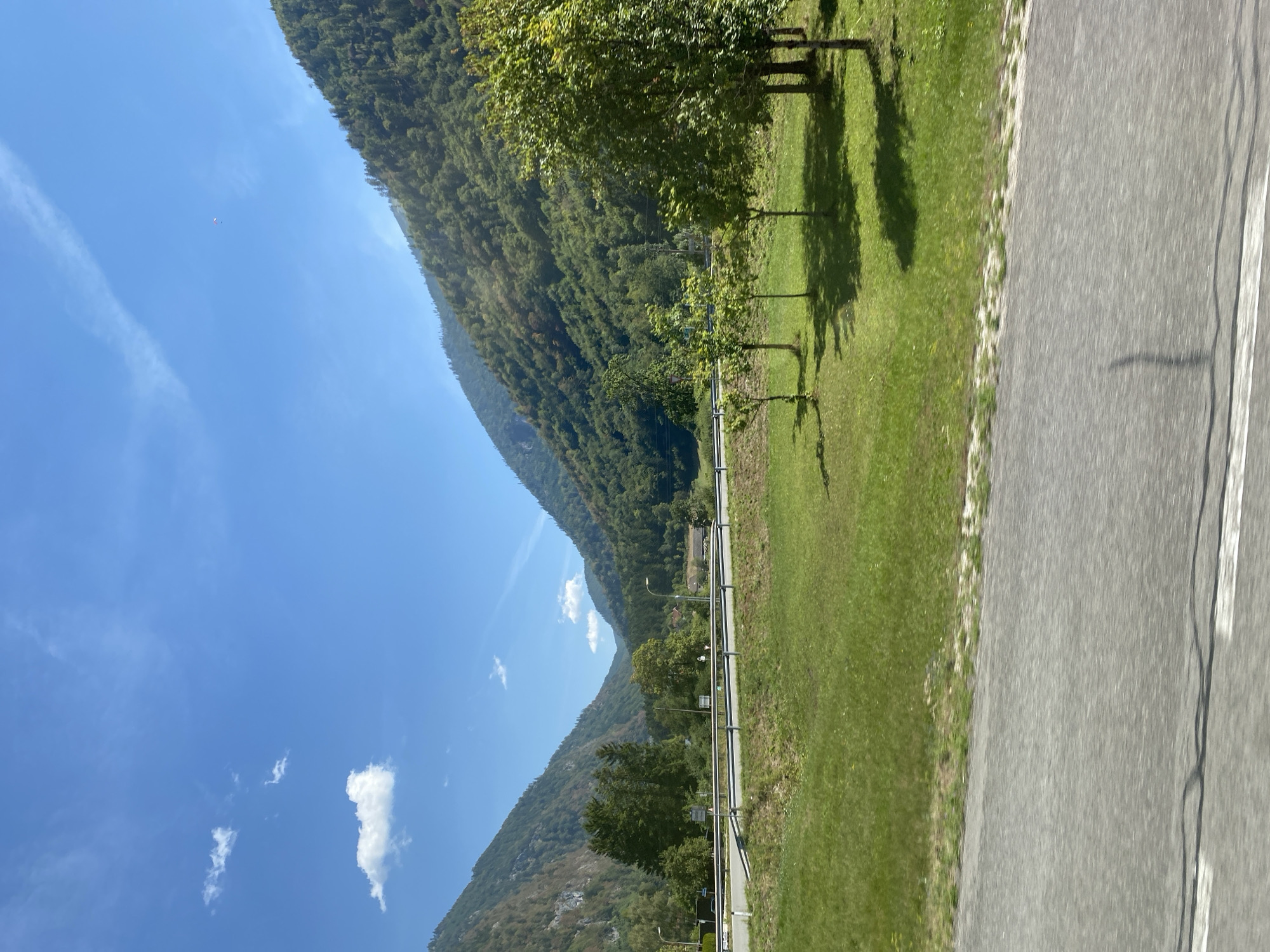 This is a photograph taken from a moving vehicle, likely on a road, looking towards a valley in Roith, Austria. The foreground shows a blurred asphalt road with white lane markings. Beyond the road, there is a grassy field with several young trees. In the middle ground, a highway or main road is visible, separated by a metal guardrail. Further back, the landscape rises into steep, tree-covered mountains on either side of a valley. The sky is a clear, bright blue with a few scattered white clouds. A small red and white object, possibly a paraglider or a kite, is visible high in the sky above the mountains. The overall impression is a scenic, rural landscape under a sunny day.