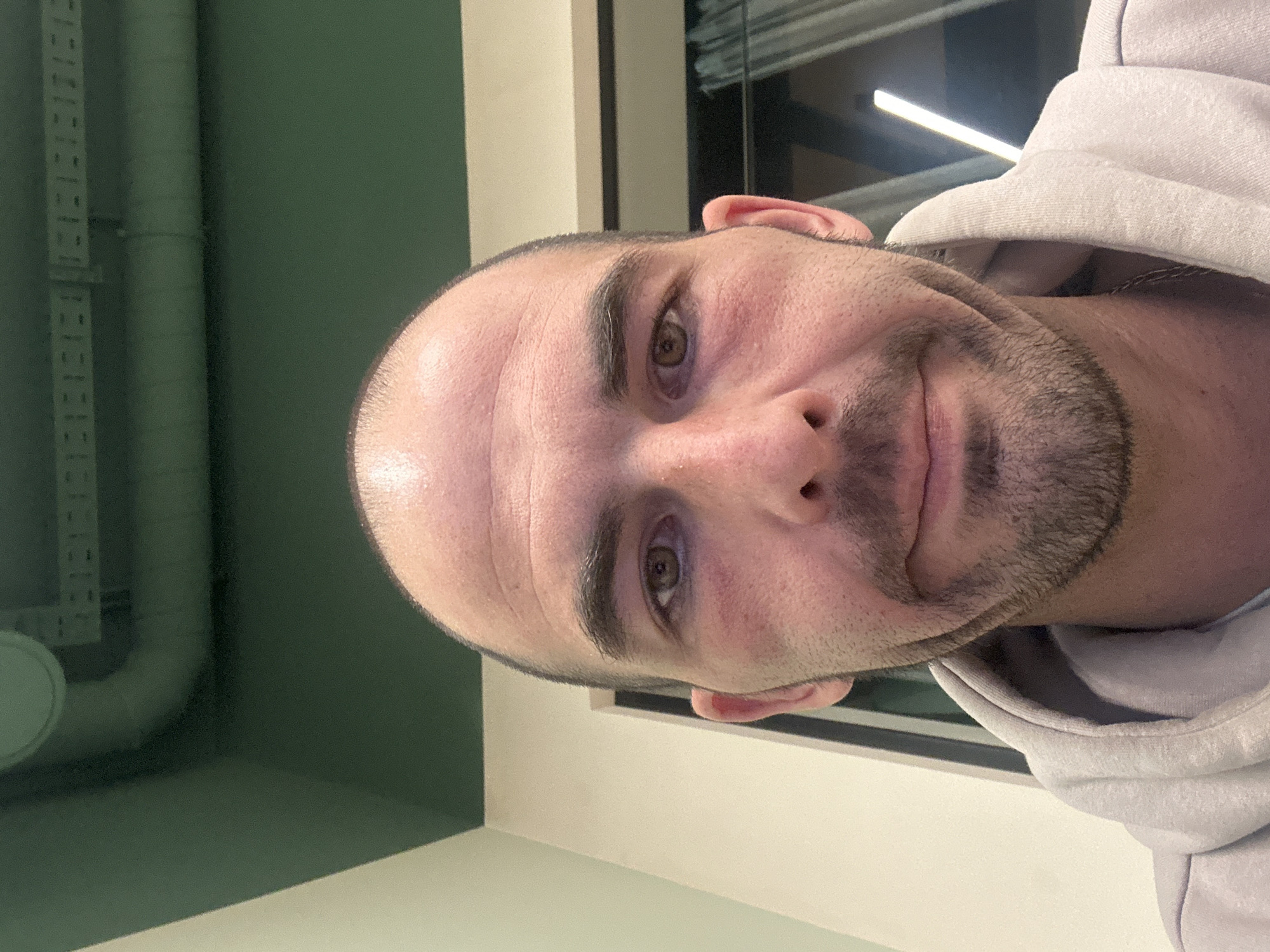 A close-up photograph depicts a man with fair skin, short dark hair and stubble, and light-colored eyes. He wears a light gray hoodie. The background consists of a green wall with pipes and a window with an interior light. There is no visual information to infer any location information, including in Budva, Montenegro.