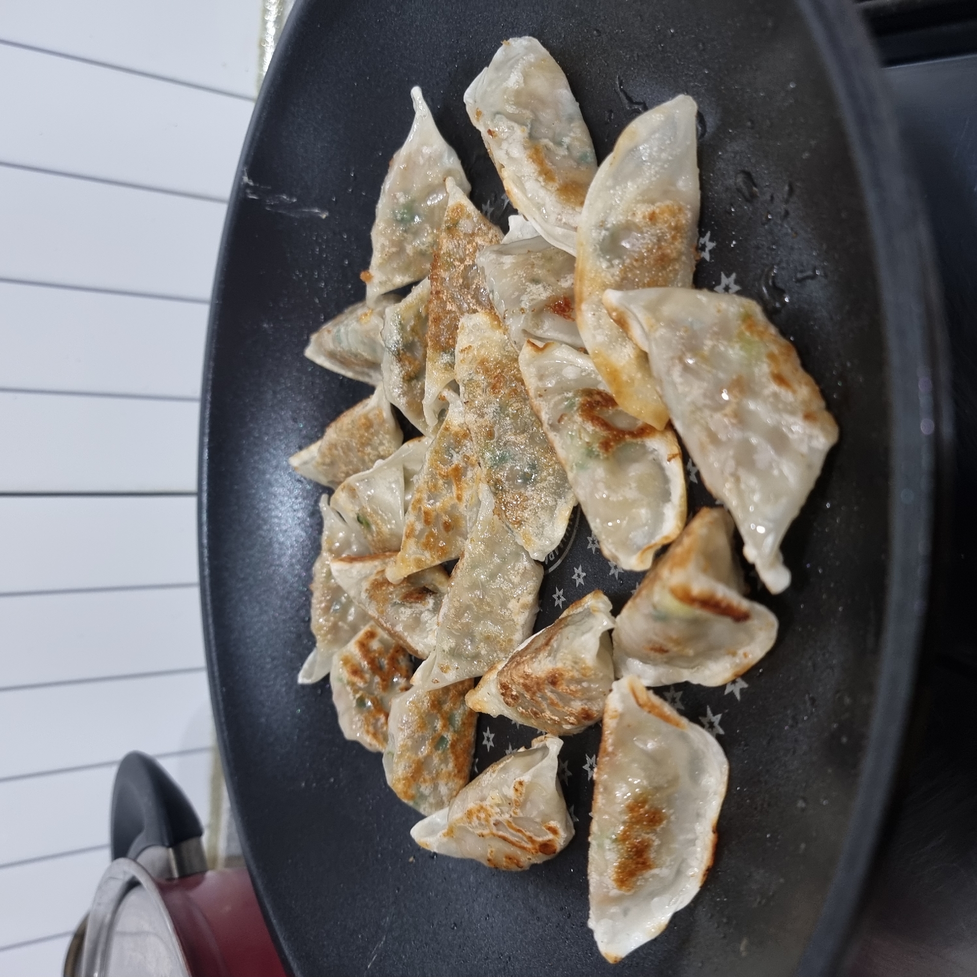 A close-up, overhead shot shows a batch of pan-fried dumplings in a black frying pan. The dumplings are arranged in a somewhat haphazard pattern, with some overlapping. They have a translucent wrapper that has turned golden brown and slightly crispy on the bottom, revealing glimpses of the filling. The scene appears to be in a kitchen, with a red pot with a silver lid visible in the upper left corner, against a tiled backsplash. The frying pan is sitting on a stovetop, and there are small droplets of oil on the pan's surface. The lighting is bright, suggesting artificial light from above. The image does not contain any people, and the dumplings are the sole focus. The location is stated as Dunnae-myeon, South Korea.