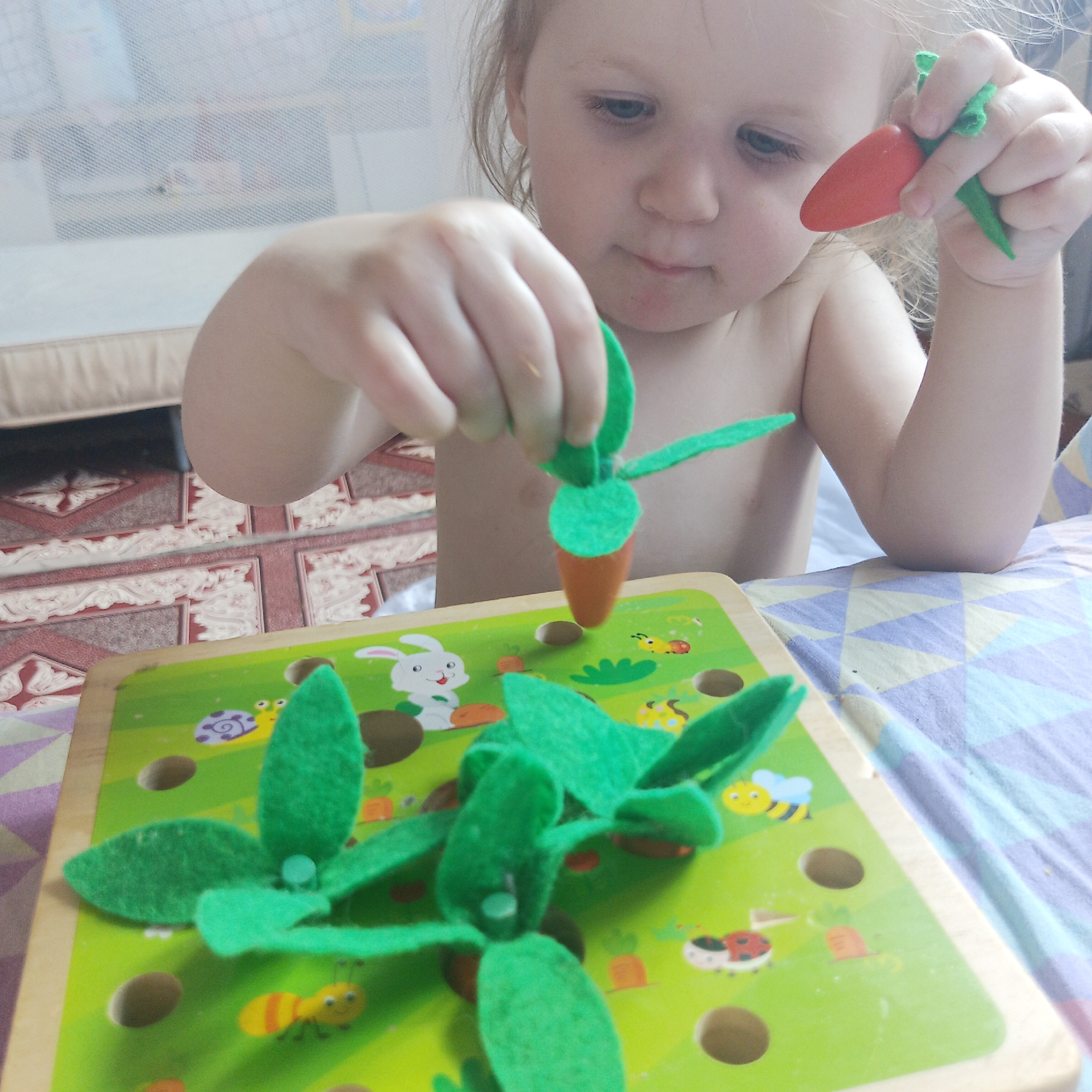 A young child with fair skin and light hair is sitting at a table, playing with a wooden toy featuring green felt leaves and orange carrots. The child holds a carrot in one hand and is in the process of inserting another carrot into one of the holes on the toy. The toy also features illustrations of a rabbit, snails, bees, and ladybugs. The child is indoors, sitting on a bed with a patterned blue and purple bedspread. A wooden crib with a net cover is behind the child. A red-patterned floor is visible in the background. The location is possibly in a residential setting in the city of Chaikovsky, Russia.