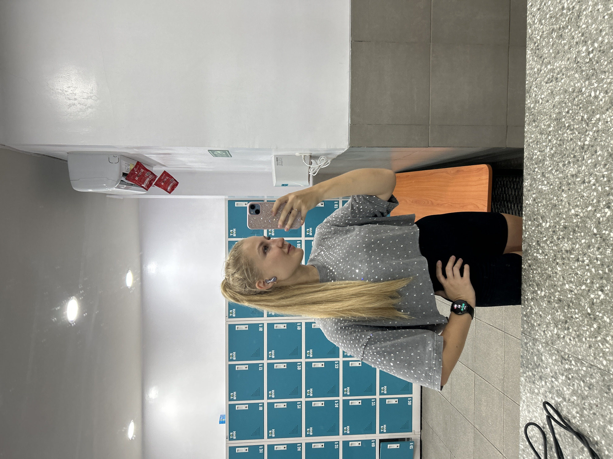 A young woman is taking a selfie in what appears to be a locker room. She has blonde hair pulled back in a ponytail and is wearing a grey, short-sleeved shirt with silver dots, black shorts, and a smartwatch on her left wrist. She is holding a pink mobile phone up to take the photo.
Behind her are rows of lockers colored in teal. The lockers have labels on them, like "HUB E.1" and number such as "E.12", "E.30", etc. Above the lockers, there is a white wall on one side and a gray tiled wall on the other. A wooden bench is visible next to the gray wall.
On the white wall, there is a white air conditioner and a white hand dryer. The room is well-lit with what appear to be overhead lights.
This image was taken indoors, likely in a gym or recreational facility. While it's impossible to determine the exact location, the locker system and environment suggest it is within a facility, not outdoors. The image does not contain any information regarding the city of Kuta Selatan, Indonesia.