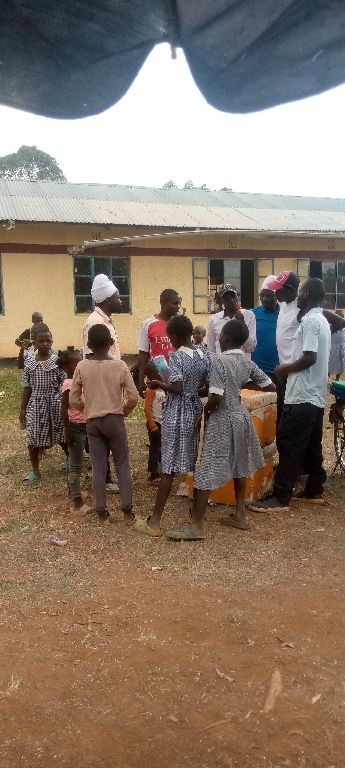 The scene is outside a building in Malava, Kenya. The building is yellow and has a corrugated metal roof. There are windows along the side of the building, and the windows are covered with window panes. The ground is dirt and there is dry grass on it. There are 12 people in the scene, including 5 adults and 7 children. The adults are wearing a variety of clothing, including shirts, pants, and hats. The children are wearing school uniforms. There are multiple objects in the scene, including an umbrella, a chair, and a water bottle. Some of the people are standing and some are sitting. The adults are talking to each other, and some of the children are listening. There is a bicycle in the background. The scene is informal and the people appear to be engaged in casual conversation. The background is out of focus, but it appears to be a rural area. The people appear to be in the process of having a meeting. The scene takes place in front of a school building, possibly during a school event.