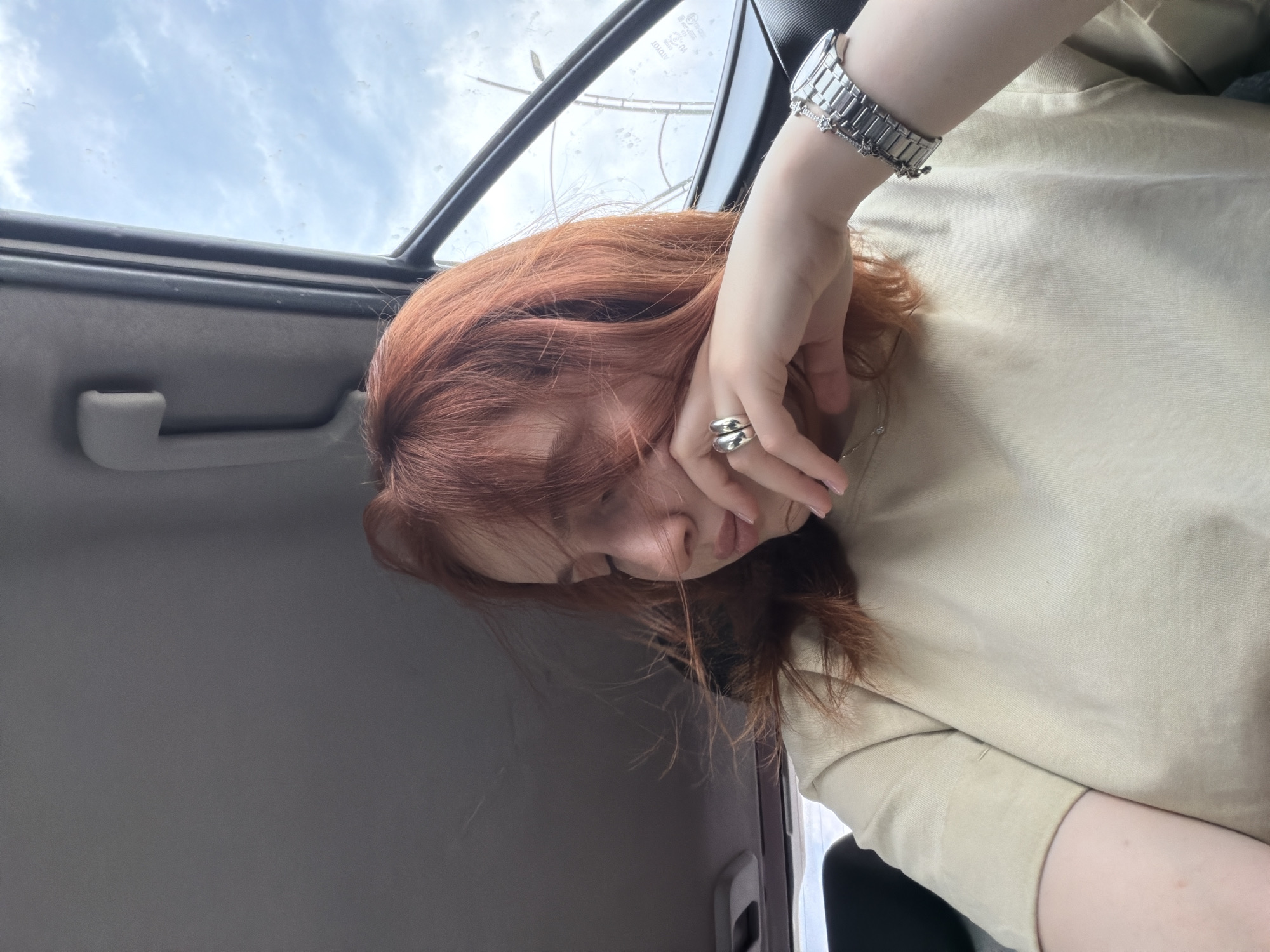 A reddish-haired person is inside a car, visible through the sunroof above, with a gray interior. The person wears a light-colored shirt and a metal watch on their left wrist. A silver ring is on one of their fingers. The location is not discernible from the image; it is not possible to identify Tbilisi, Georgia.
