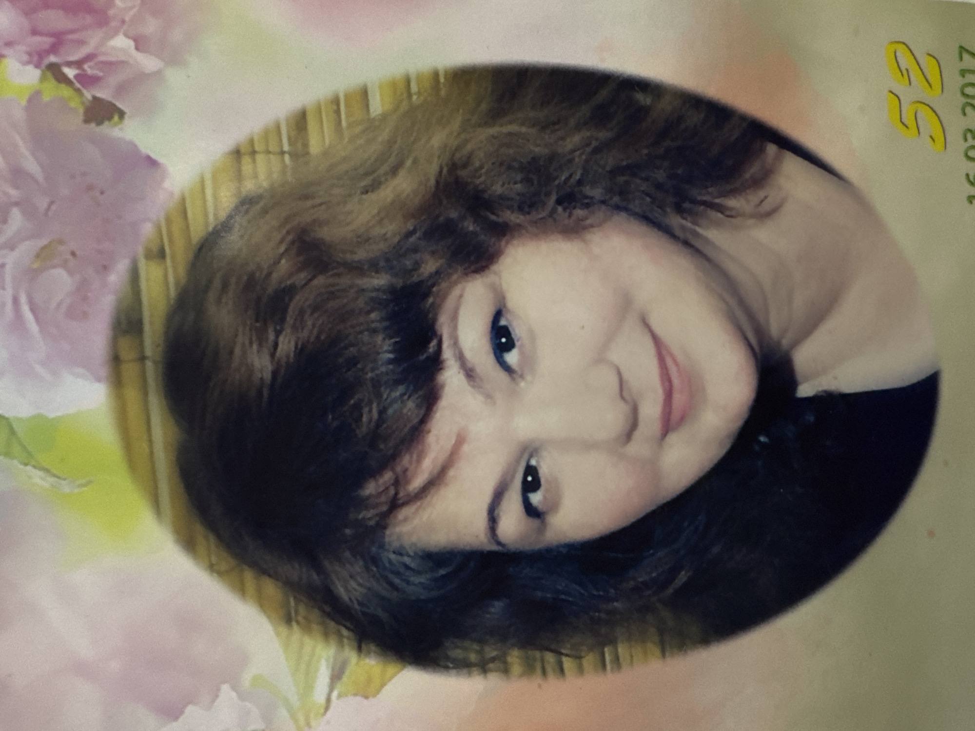 A portrait of a middle-aged woman with dark, curly hair is displayed in an oval frame. She is looking slightly to her right with a gentle smile. The background features soft, out-of-focus pink flowers and a textured bamboo-like pattern behind her head. A yellow number "52" is visible in the lower right corner of the image, along with the date "16.03.2017" below it. The overall impression is that of a memorial portrait, possibly from Izberbash, Russia, given the date and the context of the request.