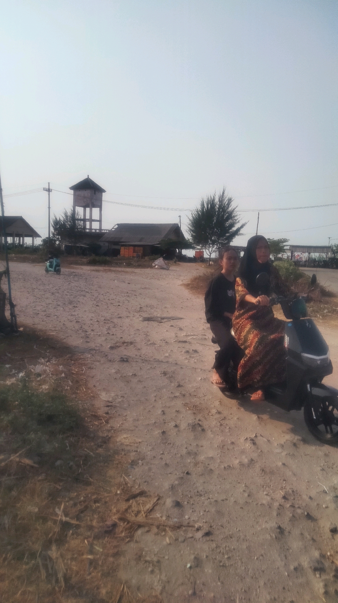 Two individuals, one in front and one behind, are seated on a dark-colored electric scooter. The person in the rear wears a patterned dress and a headscarf, while the person in front wears a dark long-sleeved shirt and dark pants. They are on a dirt road with dry grass and debris. In the background, there are several structures, including a water tower, a building with a corrugated roof, and a small, light-blue scooter. The scene appears to be in a rural or semi-rural area, consistent with locations found in Palang, Indonesia. The sky is overcast and pale.