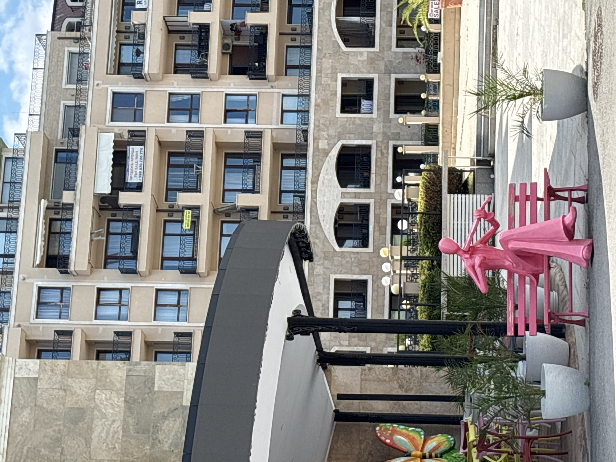 The image depicts the exterior of a modern apartment building in Varna, Bulgaria, likely in a resort area due to the presence of outdoor seating and decorative elements. The building is several stories tall with a beige facade and numerous balconies with black railings. Several windows are visible, some with curtains drawn. One balcony has a white awning and a sign advertising "SALE/RENT" in English and Bulgarian. Another balcony displays laundry.
In the foreground, a pink statue of a person playing a violin is seated on a pink bench. To the left of the statue, colorful chairs are arranged around tables, and a large, brightly colored butterfly decoration is visible. Potted plants, including palm trees and other greenery, are scattered throughout the scene. The ground is paved with light-colored tiles, and a patterned manhole cover is visible in the lower right. The sky is partly cloudy. The overall impression is one of a well-maintained, possibly commercial or residential complex in a sunny climate.