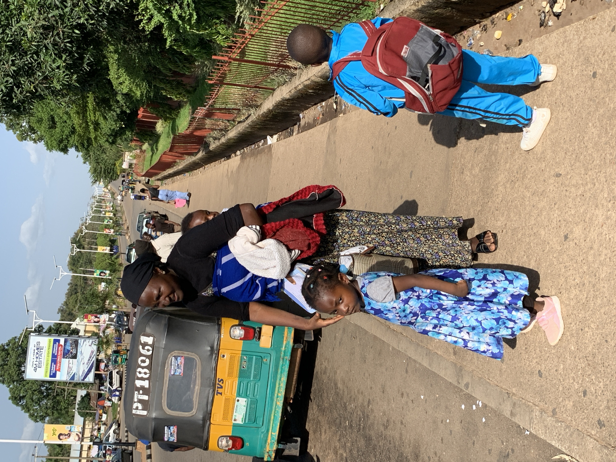 The image depicts a street scene in Jos, Nigeria, with a focus on a woman and two children on a sidewalk. The woman, wearing a black headscarf and a patterned dress, is carrying a baby wrapped in a blue cloth on her back and is holding another child's hand. The child in the foreground is a young girl wearing a blue floral dress and pink sneakers. To the right, a young boy in a blue tracksuit and white sneakers stands with a red and gray backpack. In the background, a yellow and green auto-rickshaw with the license plate "PT-18061" is visible. The street is lined with trees and has a red fence along the side. Various vehicles and a billboard are visible further down the road. The environment suggests an urban setting in Jos, likely a busy street.