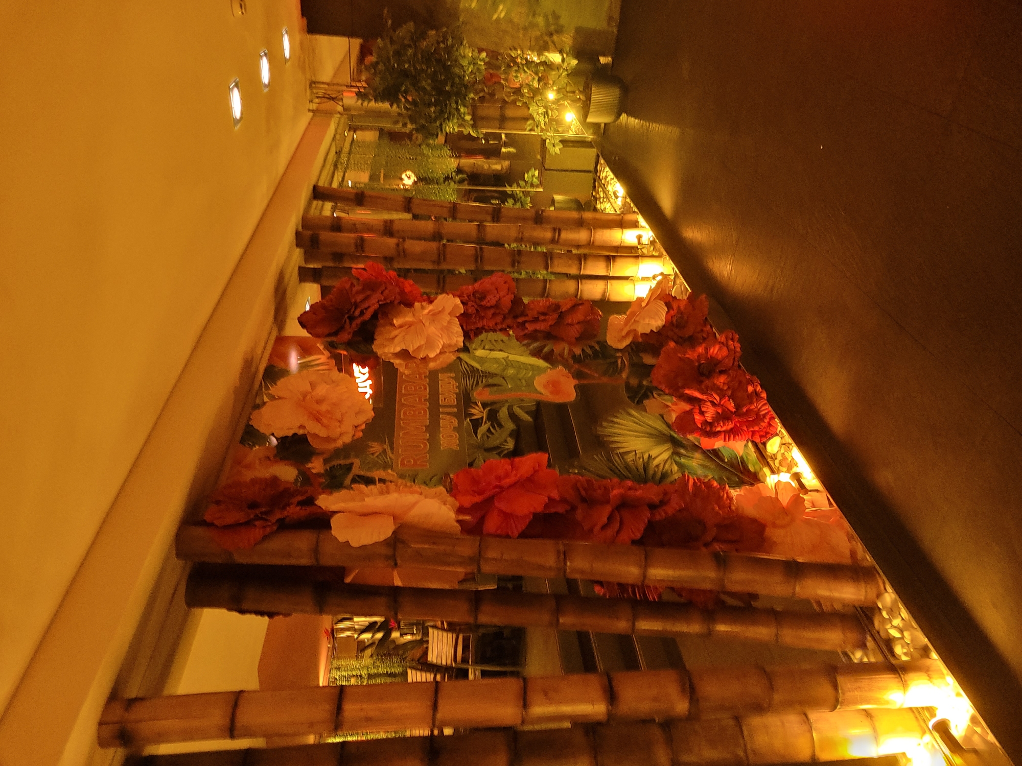 The image shows an interior walkway with a decorative bamboo wall. The wall is adorned with large, artificial flowers in shades of pink and red. Behind the bamboo and flowers, there is a glass partition that reveals a dimly lit interior space with tables and chairs. The floor is a dark, reflective material, and the ceiling is illuminated by recessed lighting fixtures. The overall ambiance is warm and inviting, with a tropical theme suggested by the bamboo and floral decorations. The text "RUMABABA" is visible on a sign behind the floral display, along with some additional text in a foreign language. The image appears to have been taken at night or in a low-light environment.