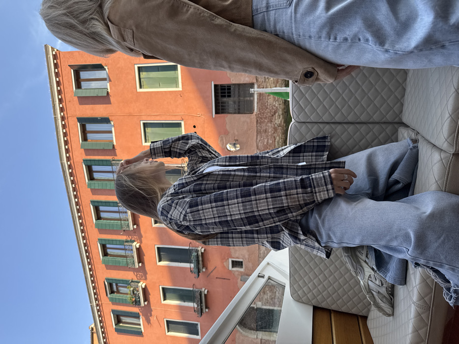 The scene depicts two individuals occupying a light-colored boat in a canal within Venice, Italy. A multi-story building with an orange-red facade dominates the background under a clear blue sky. The building exhibits multiple windows fitted with open dark green shutters; upper-floor windows include small balustrades, one containing potted plants. The building's lower section transitions to exposed reddish brickwork at the water's edge, featuring a small recessed window and a larger dark wooden door with a gridded vent.
The primary subject, positioned on the left side of the boat, is a female with long, light brown hair, seen in profile facing right. She wears a navy, beige, and white plaid shirt over a white undershirt, and light blue wide-leg jeans with distressed hems. Her right hand is raised to her forehead. A white athletic shoe rests on the quilted, light-colored upholstered boat bench near her left leg. A second individual is partially visible on the right, observed from the lower back and shoulder, wearing a tan jacket and light blue jeans, also facing right. A green and white striped mooring pole is present in the canal water between the boat and the building. Strong directional sunlight illuminates the scene.