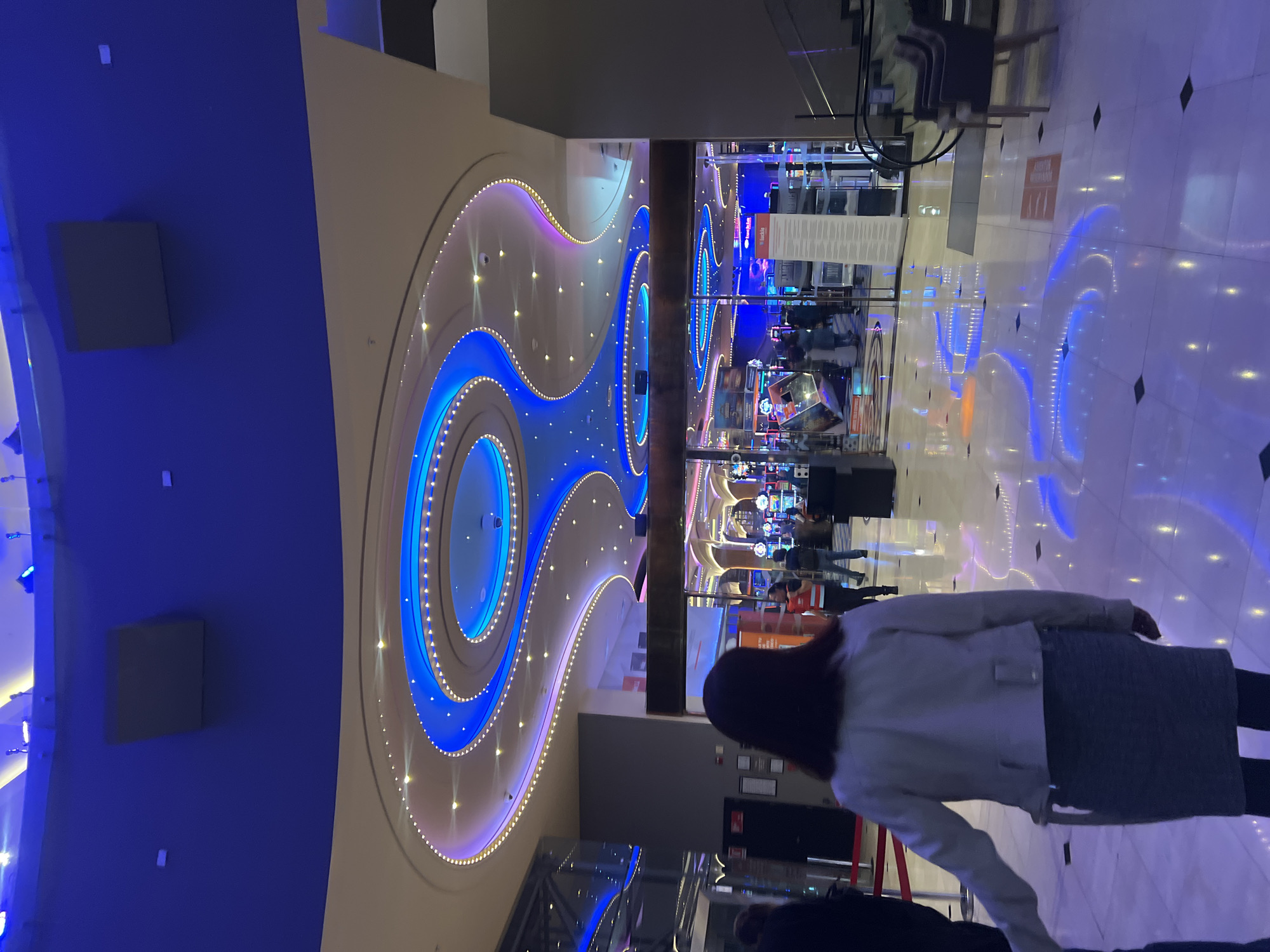 The image shows the interior of a casino, possibly in Arica, Chile, as suggested by the lighting and architectural style. The ceiling is a striking feature, decorated with swirling, illuminated blue and purple patterns. Below the ceiling, casino gaming machines and other equipment can be seen, with people milling about. In the foreground, a woman with dark hair and wearing a light-colored jacket and dark skirt is partially visible from behind, facing away from the camera and seemingly observing the casino floor. The flooring is light-colored and reflective, mirroring the ceiling lights. The overall impression is of a brightly lit, active gambling environment.