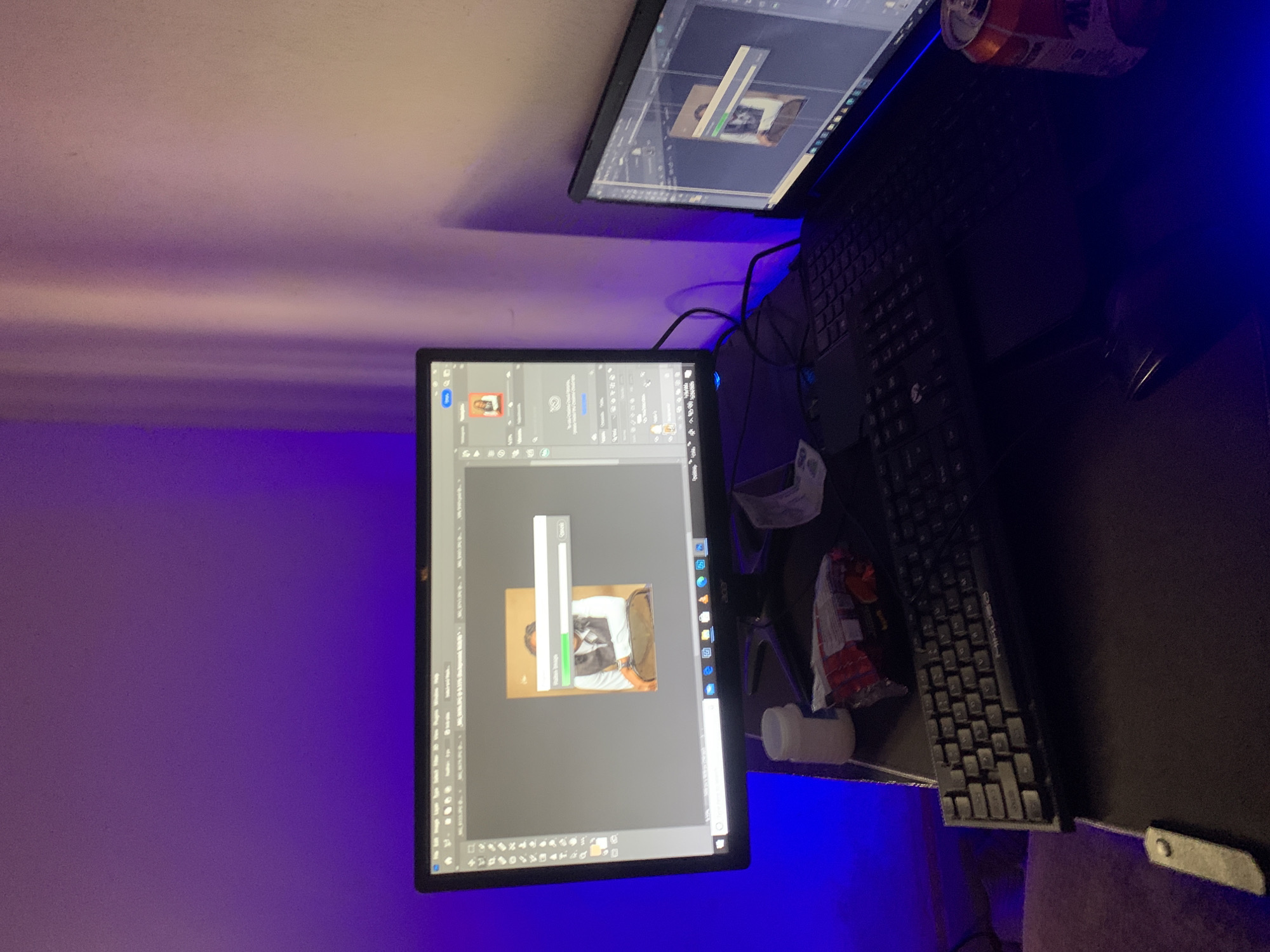 The image displays a dimly lit room with purple and blue ambient lighting. Two computer monitors are visible, one in the foreground and another slightly behind it. The foreground monitor shows a photo editing software interface, with an image of a person in a vest and shirt being edited. A progress bar is partially filled, suggesting an ongoing process. The background monitor displays a similar interface, also with an image being edited.
On the desk, a keyboard is positioned in front of the monitors. To the right of the keyboard, a soda can is precariously balanced on the edge of the desk. Various items are scattered on the desk, including what appears to be a small white container and a crumpled plastic bag. The overall setting appears to be a home office or workspace. The time of day is not discernible, and there are no weather cues. The image provides no specific location cues that would indicate Gwafan, Nigeria, beyond what is stated in the prompt. There is no visible text in the image that provides additional information.