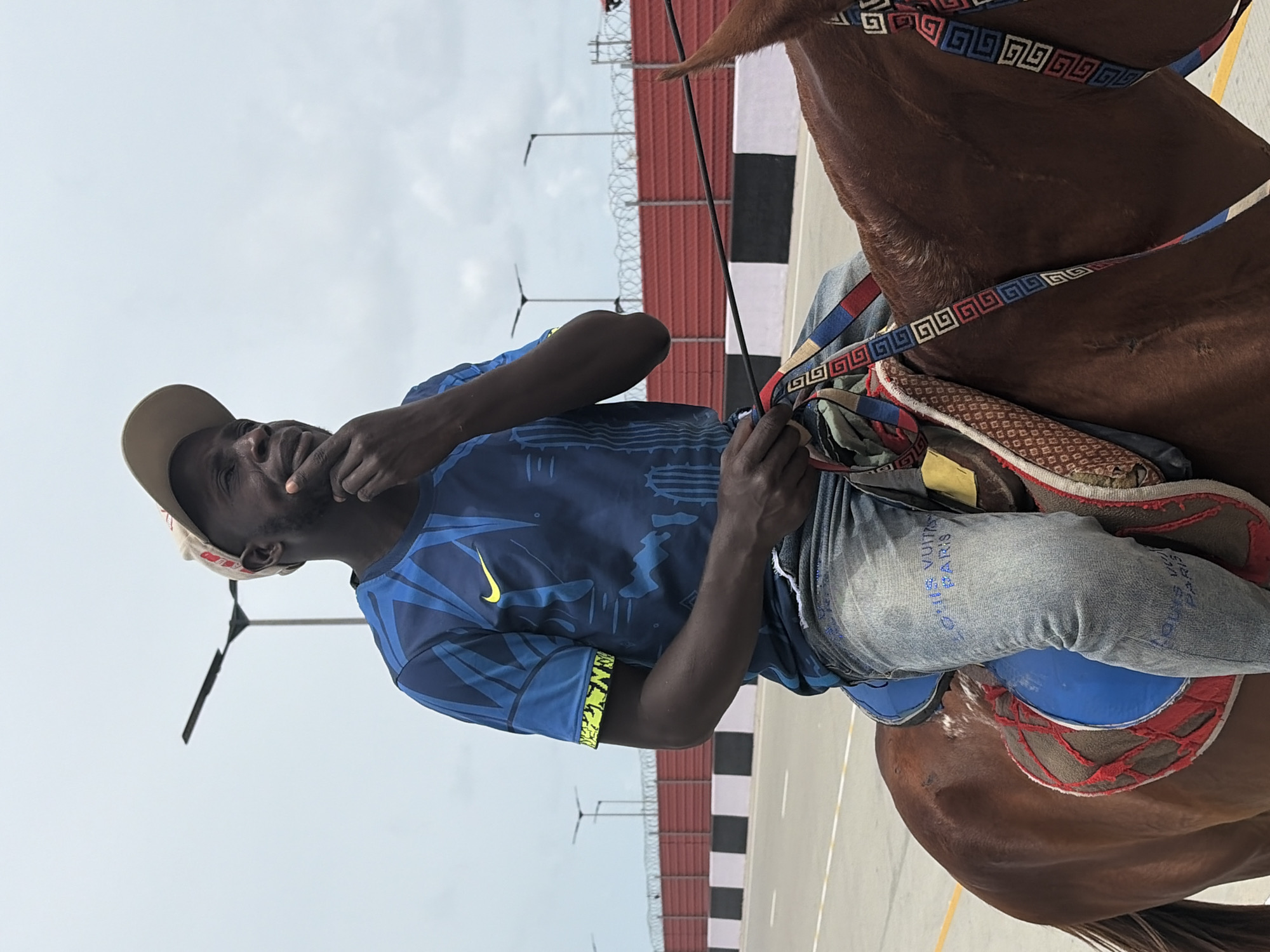 A man wearing a tan baseball cap, blue shirt with a yellow Nike logo and blue patterned sleeves, and light blue jeans with "LOUIS VUITTON PARIS" printed on them, is sitting atop a brown horse. The horse has decorative straps and a saddle. The man is holding a black stick and has his hand near his mouth. In the background is a red fence, lampposts, and a light gray sky. The location appears to be an outdoor area in the city of Alaguntan, Nigeria.