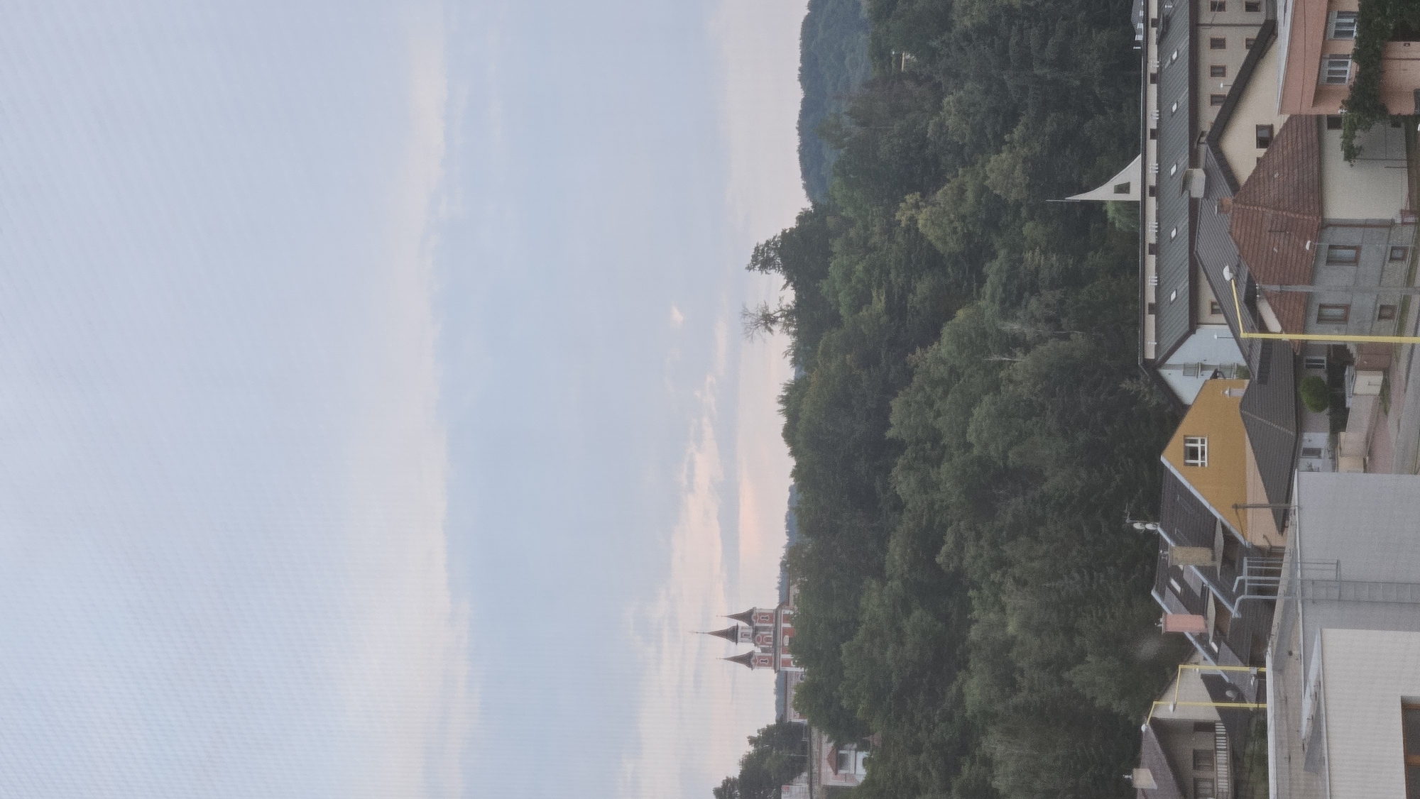 The image depicts a panoramic view of a residential area in Prešov, Slovakia, with a prominent red and white building resembling a church or monastery situated atop a densely forested hill in the background. The sky is overcast with soft, muted colors, suggesting it is either early morning or late evening. In the foreground, several houses with diverse roof styles and colors are visible, indicative of a typical urban or suburban landscape. The architecture suggests a mix of modern and older structures. The overall scene is tranquil and appears to be a quiet residential neighborhood.