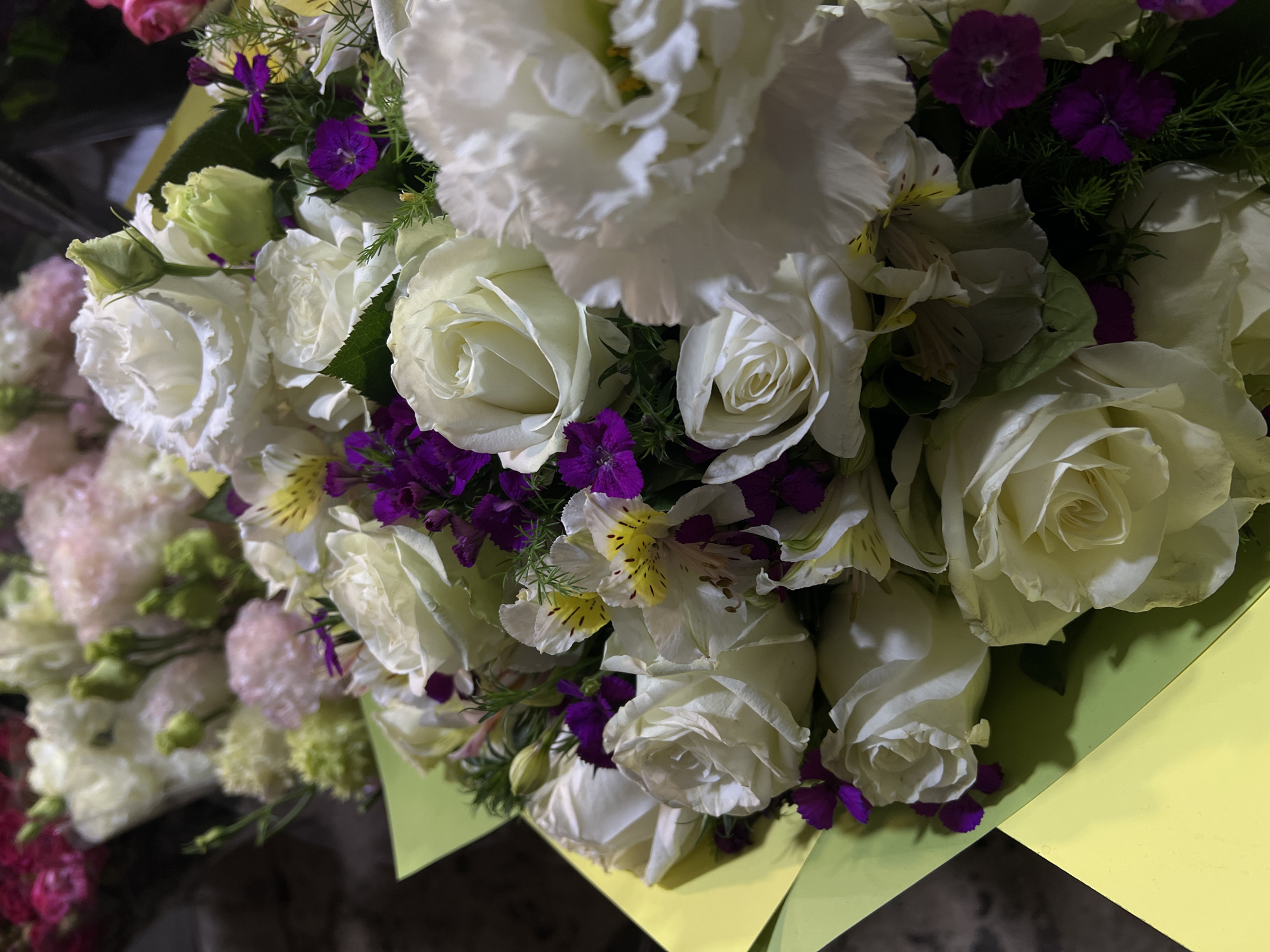 This image depicts a bouquet of white roses and other flowers, including purple dianthus and white alstroemeria. The bouquet is wrapped in yellow paper and is likely indoors, given the lighting. The location is unknown.