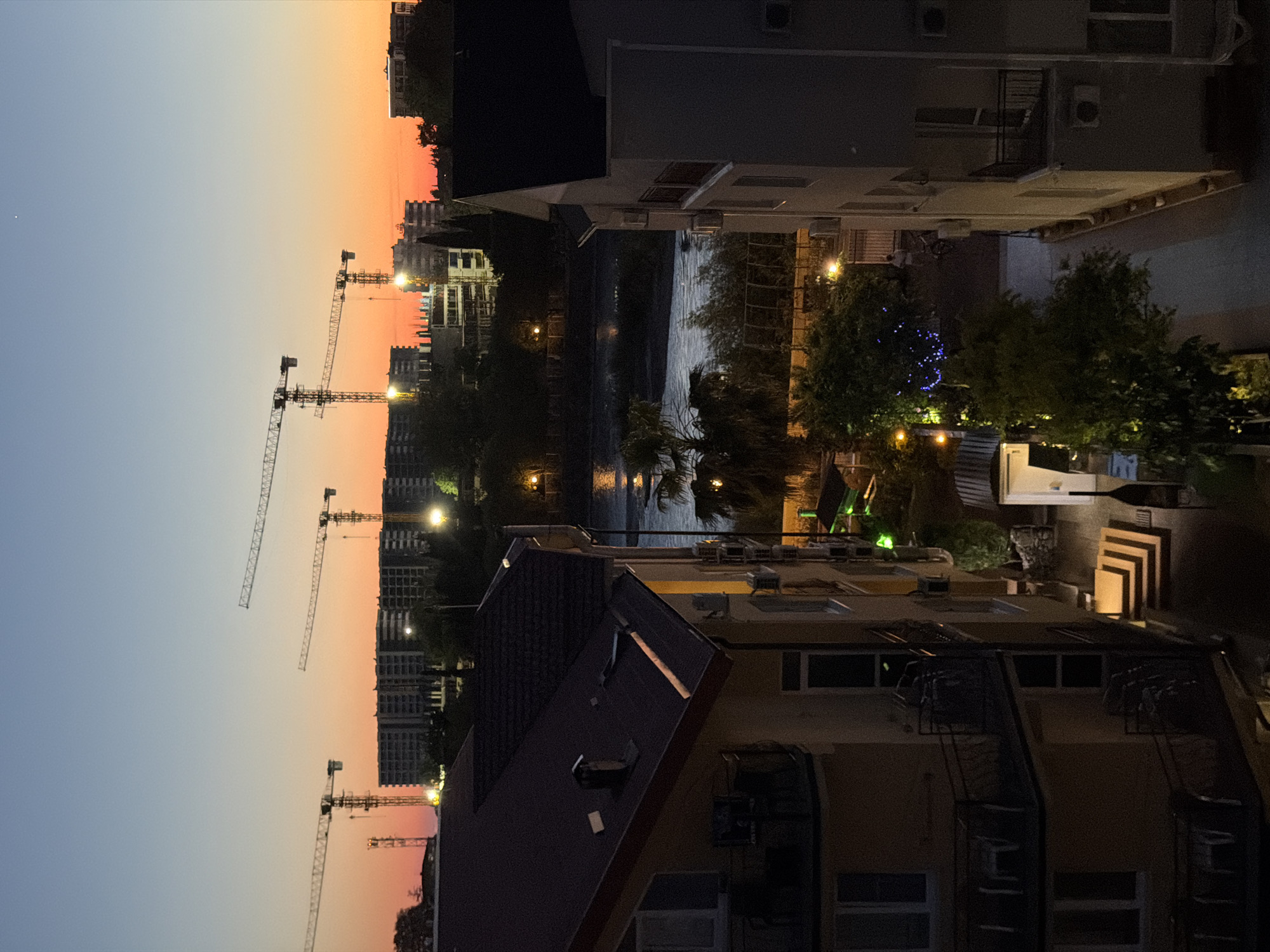 This is a long shot photograph taken from an elevated perspective at dusk in Sirius, Russia, likely overlooking a developing urban area. The sky displays a gradient of colors, transitioning from a pale blue at the top to a warm orange and pink near the horizon. Dominating the background are several tall construction cranes, silhouetted against the colorful sky, indicating ongoing building projects. In front of the cranes, incomplete multi-story buildings with exposed concrete structures are visible, suggesting a construction site.
Closer to the foreground, several residential buildings are present. On the left, a multi-story building with dark-tiled roofs and balconies is partially visible. On the right, another building, primarily rendered in muted grey tones, also features balconies and appears to be residential. Between these buildings, a body of water, possibly a river or canal, is visible, reflecting the ambient light. Trees and vegetation are scattered throughout the scene, some illuminated by streetlights.
In the middle ground, between the residential buildings and the construction site, a pathway or courtyard area is visible. There are stairs leading down to this area, illuminated by artificial light, and a few pieces of outdoor furniture on balconies. Further back, near the water, a palm tree is noticeable. The overall impression is one of a city undergoing development, with both completed and under-construction structures coexisting with natural elements. The lighting suggests the transition from daylight to night, with artificial lights beginning to illuminate the scene. There are no discernible individuals or specific actions captured in the image.
