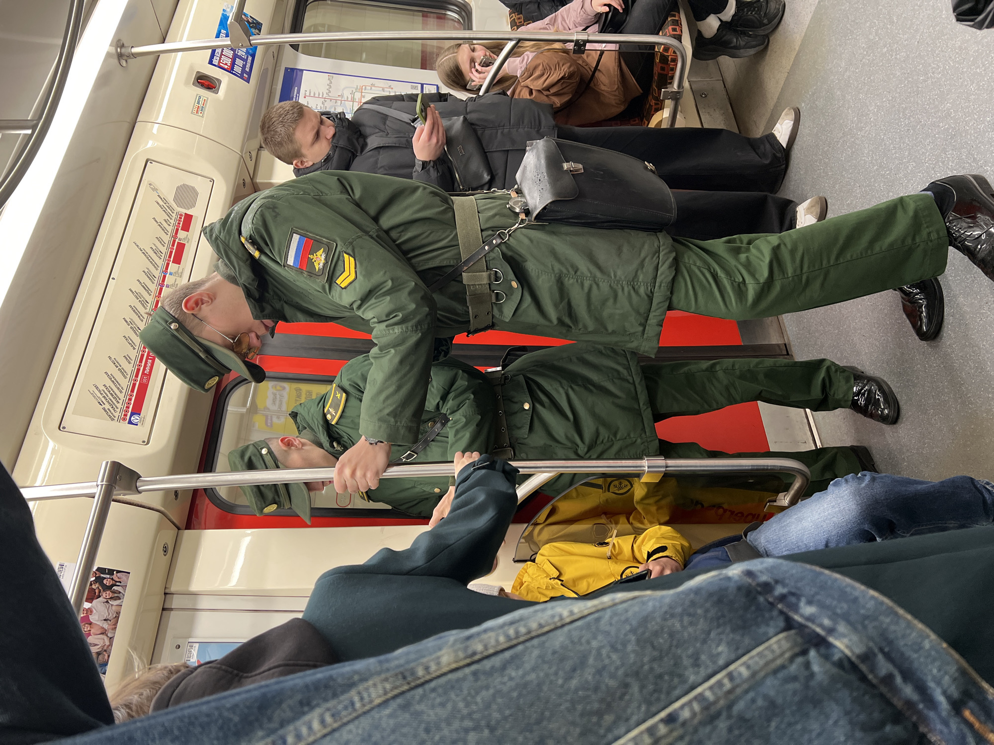 The image shows the interior of a train, likely in Saint Petersburg, Russia, judging by the Russian subway map visible. There are several people present. A person in what appears to be a military-style green uniform with a Russian flag patch is standing and holding onto a metal bar. To the side of this person is another individual in a black jacket and pants. In the foreground are the legs and lap of a person wearing blue jeans and a dark blue jacket. Also visible in the background are other passengers, including one person wearing a bright yellow jacket, partially obscured by the foreground. The setting is a public transit environment, likely a subway car during transit.