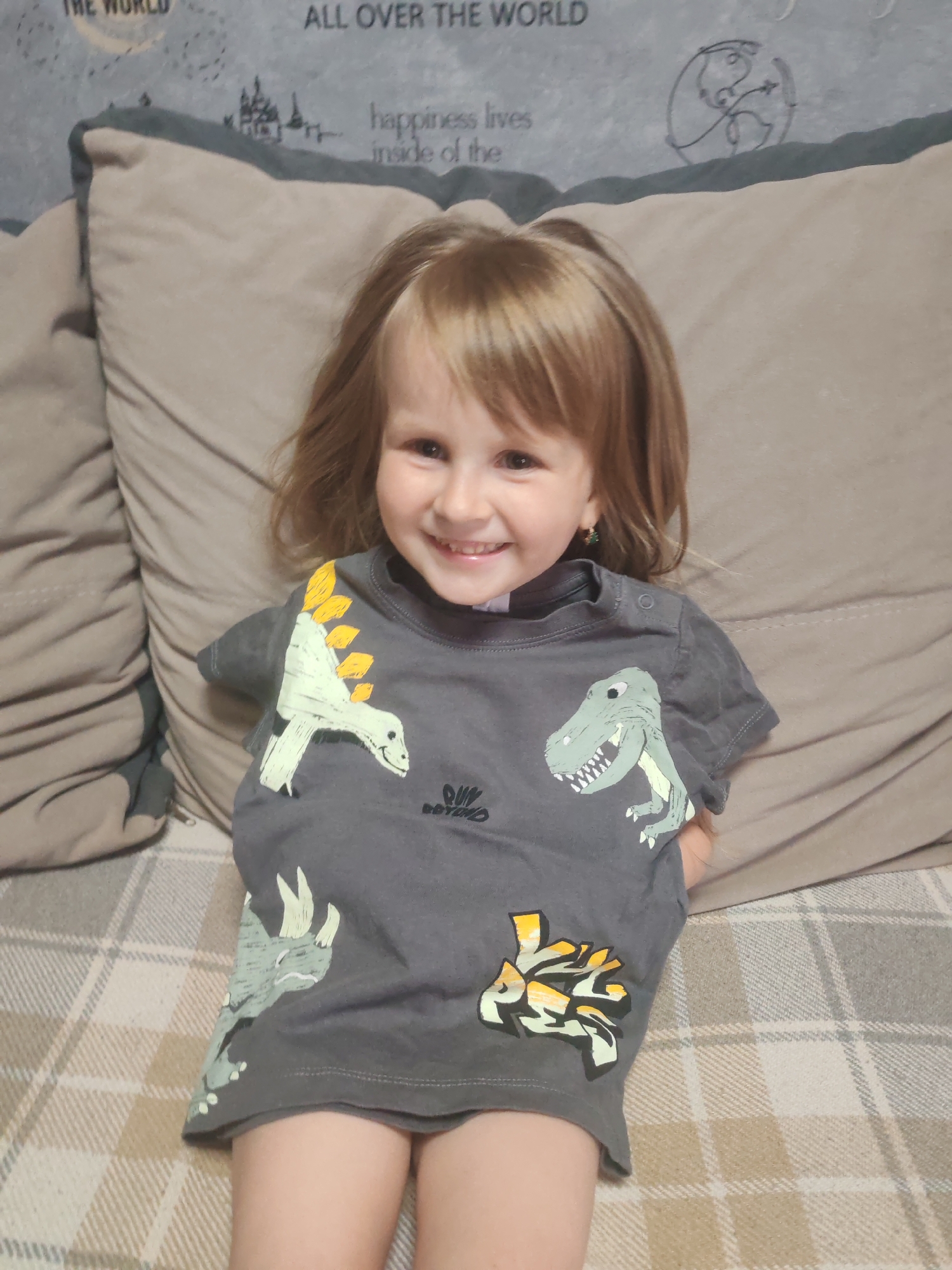 A young girl with blonde hair and a bright smile is sitting on a couch. She is wearing a dark gray t-shirt with various dinosaur graphics. The couch has large, beige pillows and a plaid blanket is visible on the seat. The background shows a wall with decorative text and images. The lighting suggests it is daytime indoors. The girl appears happy and playful. Visible text on the wall includes "ALL OVER THE WORLD" and "happiness lives inside of the". There is also a logo on the girl's t-shirt. The image was likely taken in Mezhdurechensk, Russia.