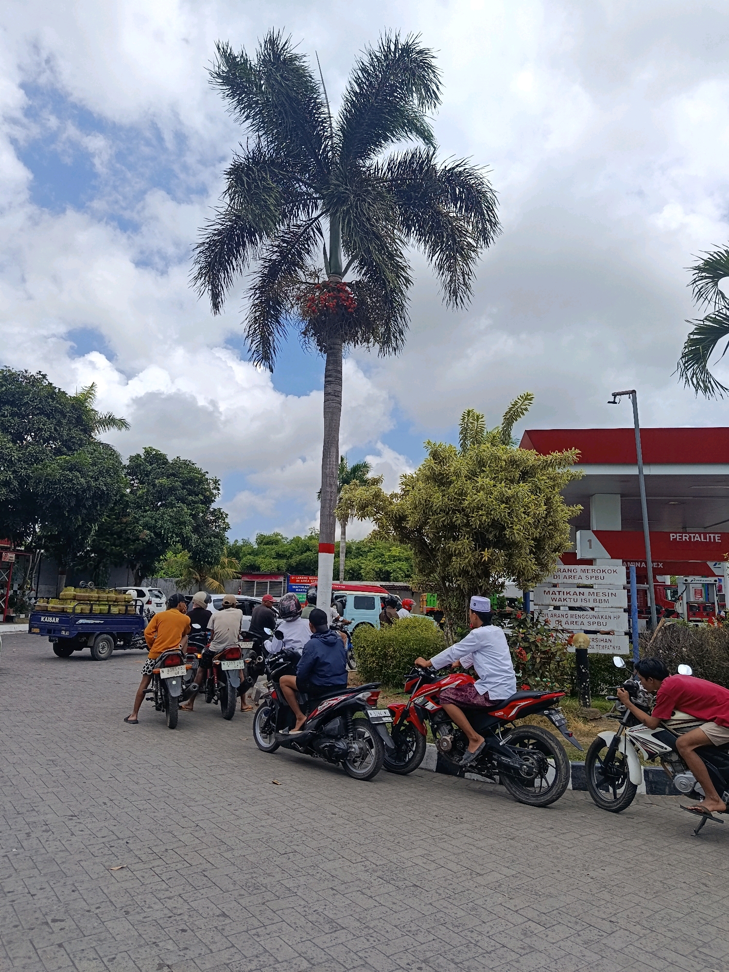 The image depicts a scene at a Pertamina gas station in Tempeh Lor, Indonesia. In the foreground, several people are on motorcycles, some waiting in line. A blue truck is parked on the left side of the image, loaded with what appear to be large green fruits, possibly durian. Palm trees and other greenery are visible, with a large palm tree with red fruit clusters dominating the upper half of the frame. The sky is partly cloudy with a bright blue sky visible. A red and white Pertamina station canopy is in the background. Signs with Indonesian text are posted near the gas pumps. The ground is paved with interlocking bricks.