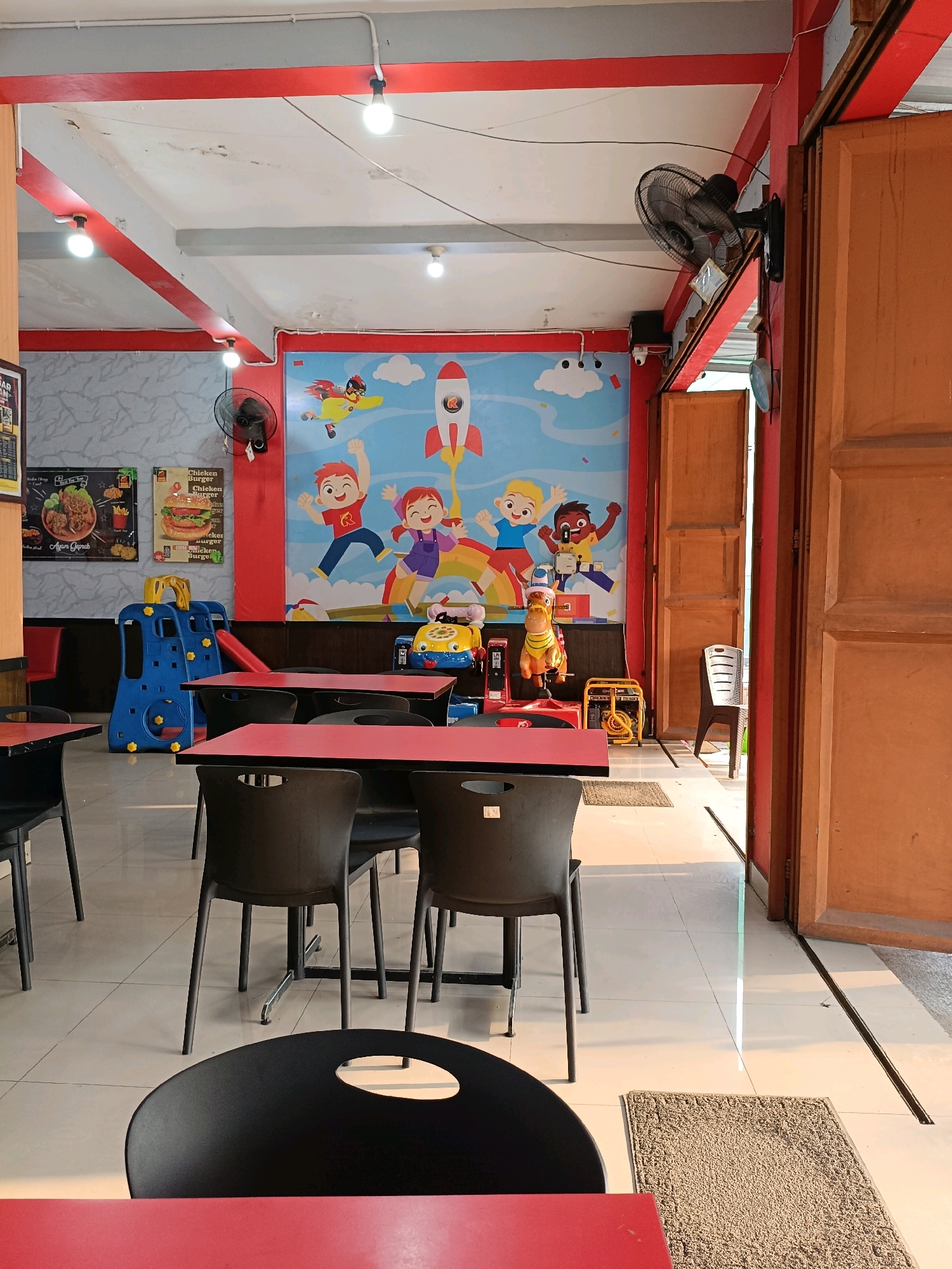 The image depicts the interior of a brightly lit restaurant, likely a family-friendly establishment, located in Ngemplak, Indonesia. The walls are adorned with a vibrant mural featuring cartoon characters flying on rockets and playing, suggesting a children's theme. Red and grey beams crisscross the ceiling, supporting fluorescent light fixtures.
In the foreground, a red table is partially visible, with a black chair in front of it. Further back, several black chairs are arranged around red-topped tables. To the left, a blue and yellow plastic play structure with a slide is set up, indicating a play area for children. Several posters advertising food, possibly chicken dishes and burgers, are hung on the wall.
A black oscillating fan is mounted on the wall, and a small security camera is also visible. To the right, a large wooden double door stands ajar, revealing a sliver of the outside. The floor is tiled with a light-colored square pattern.
The overall impression is of a clean, casual dining space designed to appeal to families with young children. The lighting is bright, and the decor is cheerful and engaging.