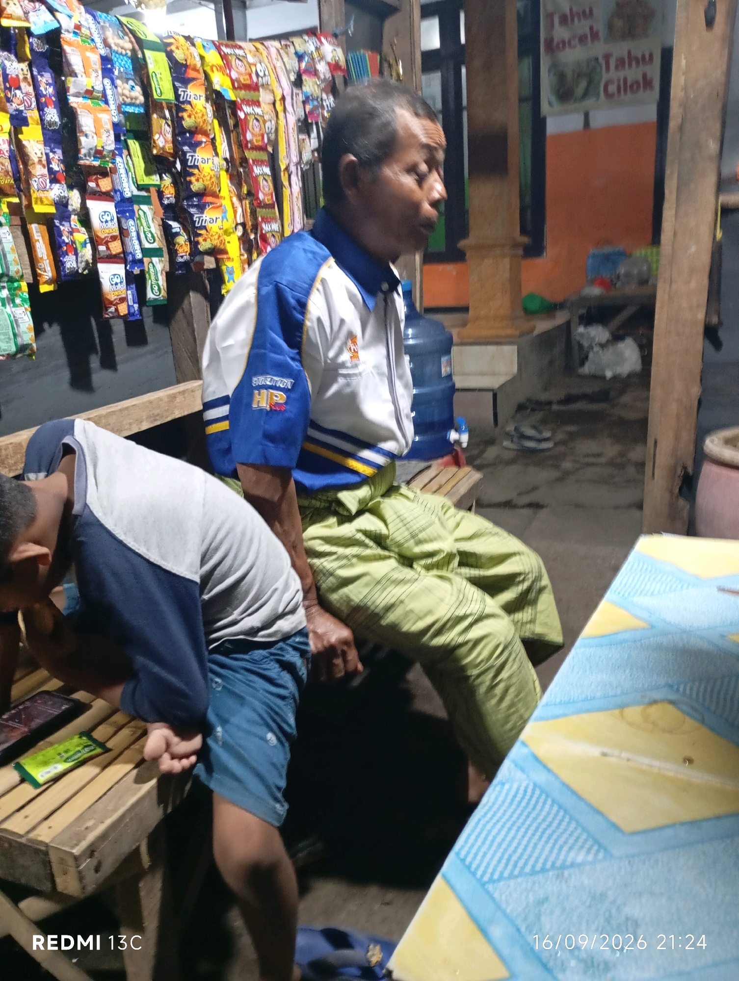 In Wuluhan, Indonesia, a man in a blue and white shirt and green pants sits on a wooden bench, facing right. His left hand rests on his leg, and his right hand is out of frame. Beside him, a younger person in a grey and blue shirt and blue shorts bends over, their hands near their lap, looking down. They are seated on a similar wooden bench. In the background, a shelf displays rows of colorful snack packets. A blue water cooler stands to the left of the older man. The environment appears to be an outdoor stall or shop, with a wooden pillar and a wall visible behind the people. A table covered with a blue and yellow patterned cloth is in the foreground. The timestamp "16/09/2026 21:24" is present in the lower right corner.
