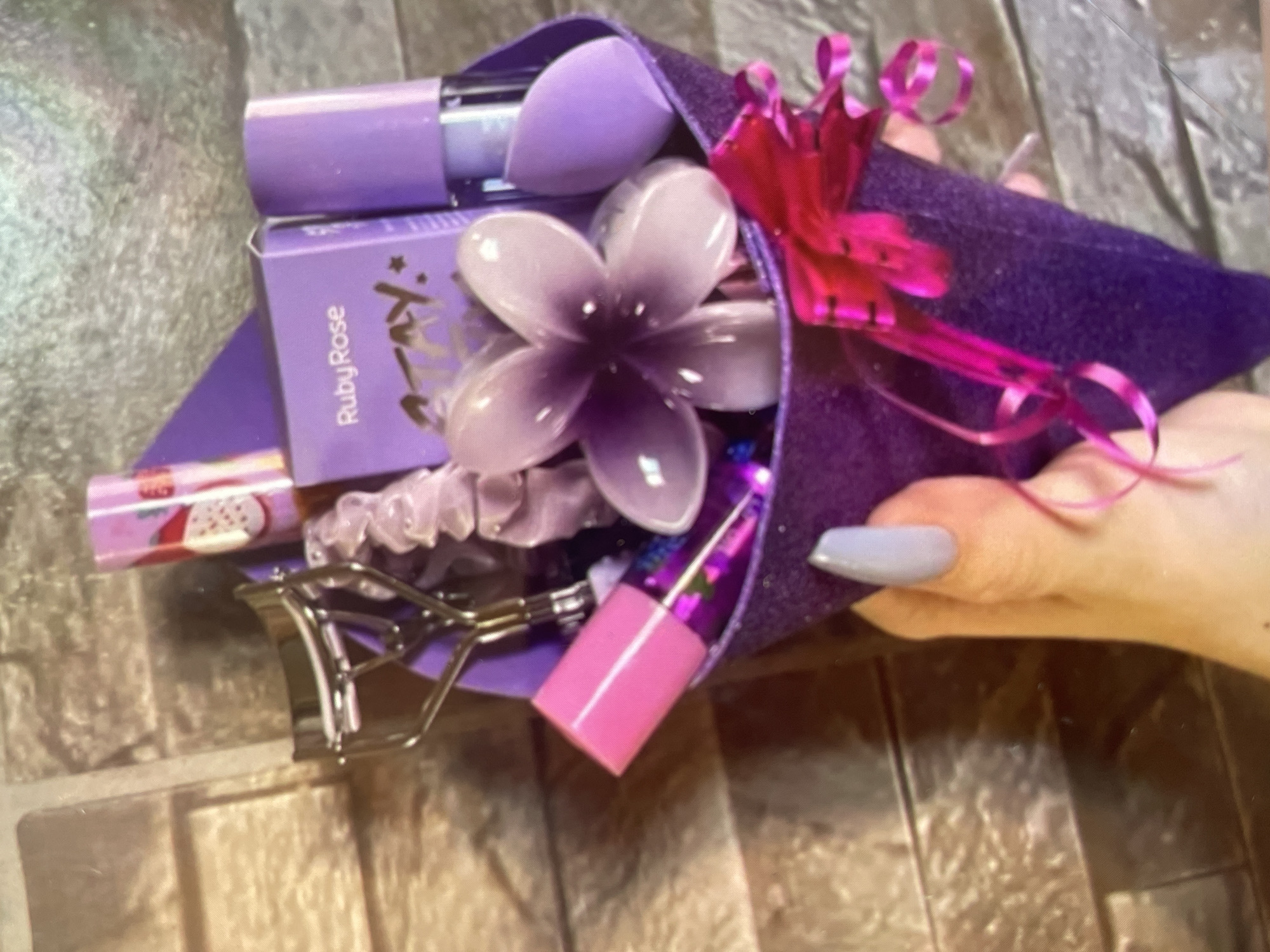A hand with a polished light-blue fingernail holds a purple cone filled with various cosmetics. The cone is decorated with a pink ribbon and a large artificial flower. Visible items inside the cone include a makeup blender, a Ruby Rose product box, a lipstick, a hair tie, and an eyelash curler. The background appears to be a tiled surface. The location is not discernible from this image.