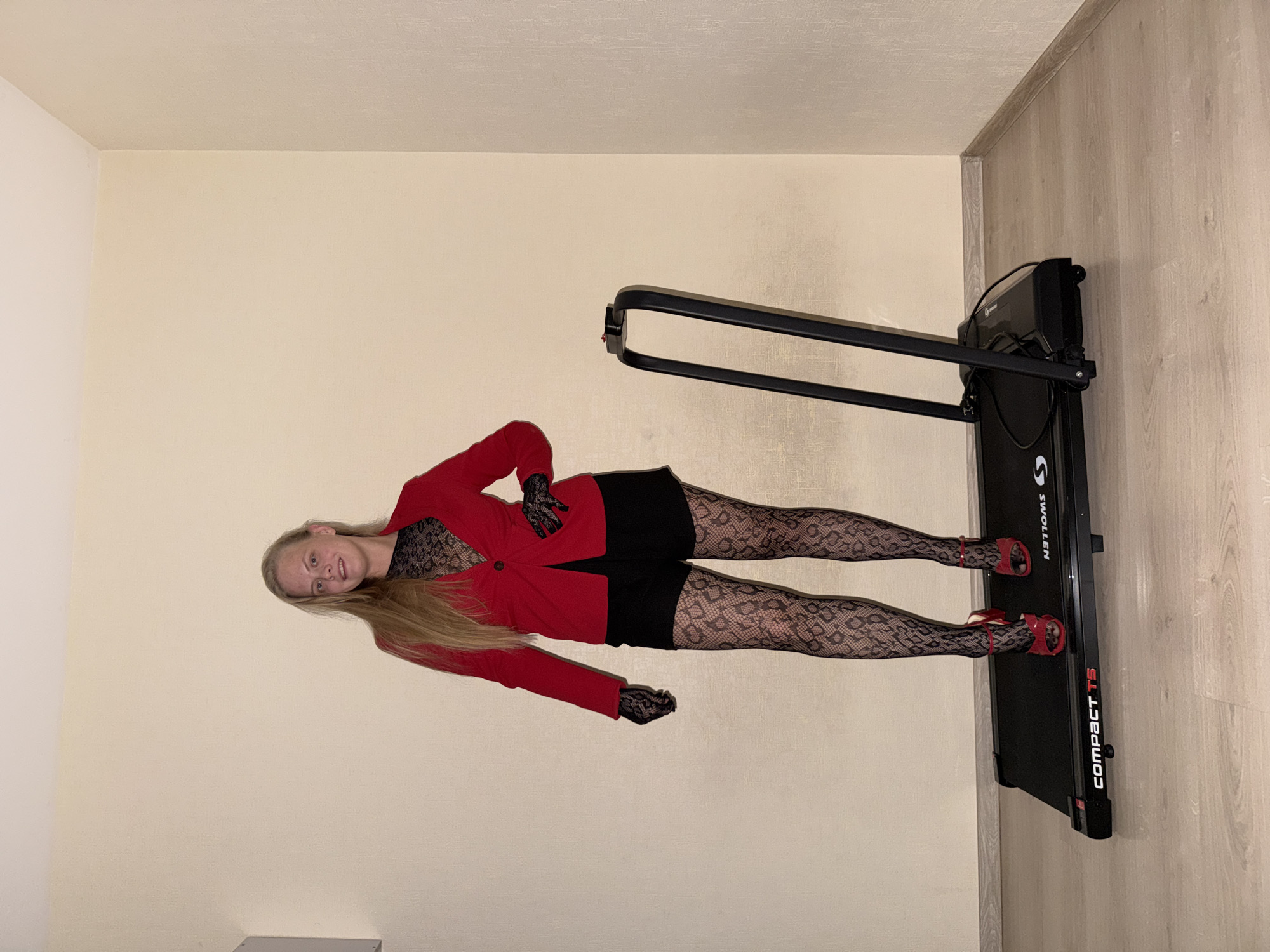 A woman with long blonde hair stands in an indoor room, posing next to or on a black treadmill. She is smiling subtly and looking towards the camera. The setting appears to be a modern interior in Izhevsk, Russia, with light-colored walls and light wood-look laminate flooring.
The woman is dressed in a vibrant red blazer or jacket layered over a black lace top or bodysuit. She wears black shorts, intricate black patterned hosiery or tights, and red high-heeled sandals. Black lace fingerless gloves complete her ensemble. Her posture is a relaxed pose, with one hand on her hip.
The treadmill is black and has white and red branding visible on its side. The text "SWOLLEN" is displayed prominently, along with "COMPACT T5". The room is lit with artificial indoor lighting. The scene suggests a staged photoshoot, emphasizing the woman's attire.