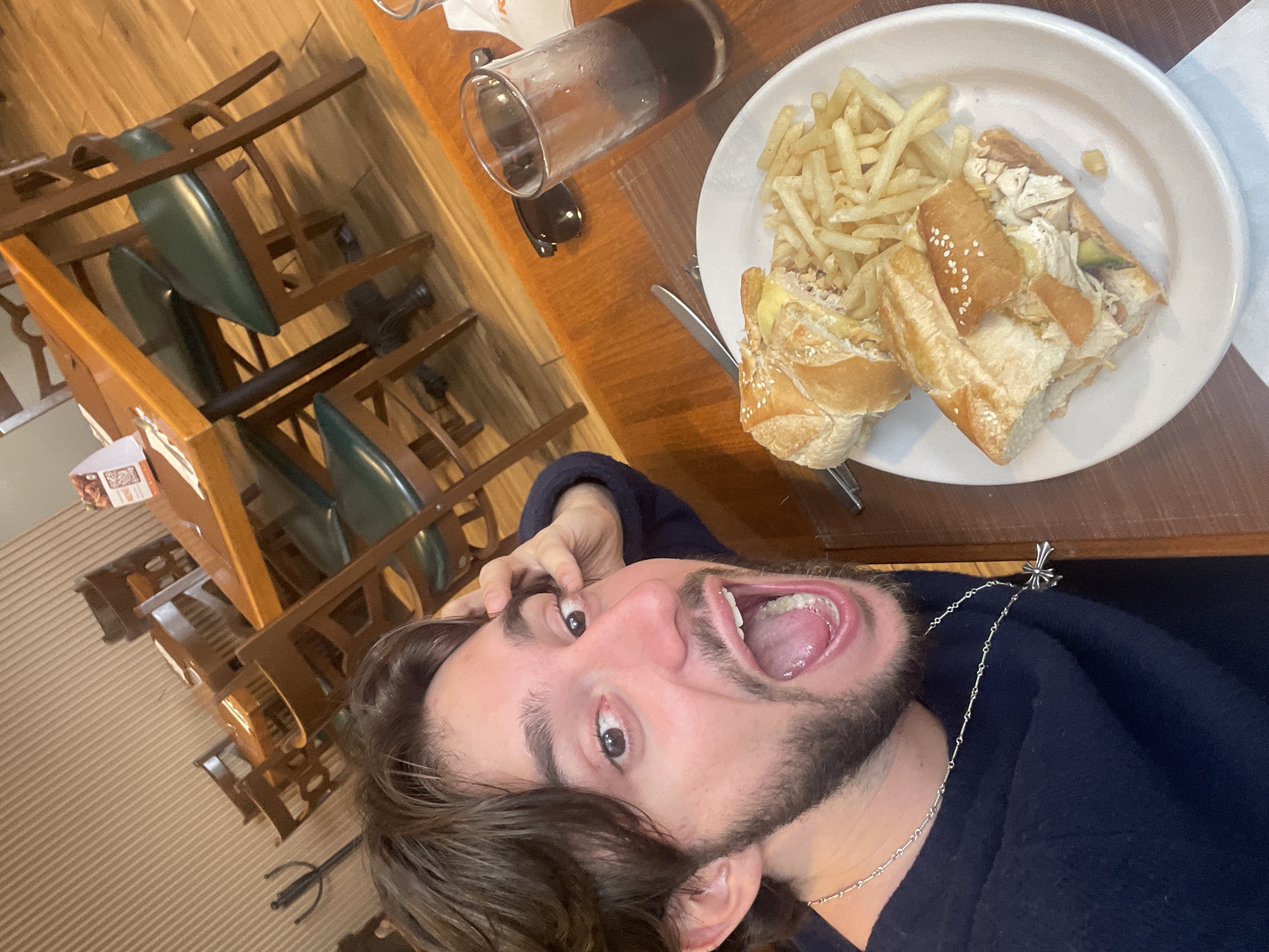 A young man with dark, medium-length hair and a beard is taking a selfie in a restaurant. He is wearing a dark blue sweater and a silver cross necklace. His mouth is open in a surprised expression, and he is touching his head with his right hand. In front of him on a wooden table is a white plate with a sandwich cut in half and a pile of french fries. To the right of the plate is a glass of dark liquid, likely soda, and a pair of dark sunglasses. The background shows a dining area with wooden tables and chairs, and a wall with horizontal wood paneling. The restaurant appears to be located in Cuautitlán Izcalli, Mexico.