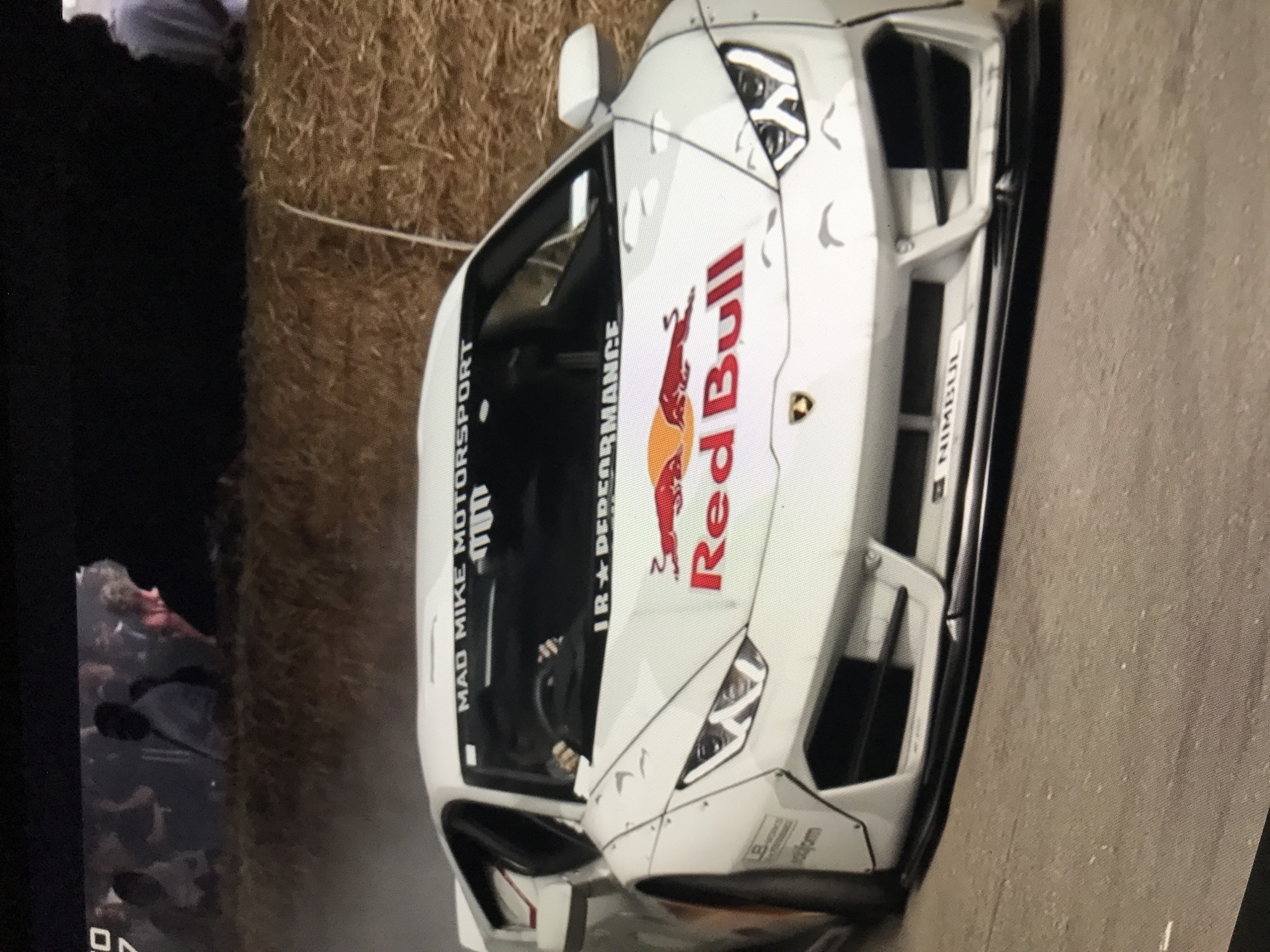 The image shows a white Lamborghini racing car with Red Bull and "Mad Mike Motorsport" branding. It appears to be situated on a course or track, as evidenced by the bales of hay. There are people visible in the background, suggesting a spectator area. Based on the license plate "NIKISUL", the location can be deduced as Donji Pobori, Montenegro. The car is angled towards the upper right, and the overall impression is of a racing event.