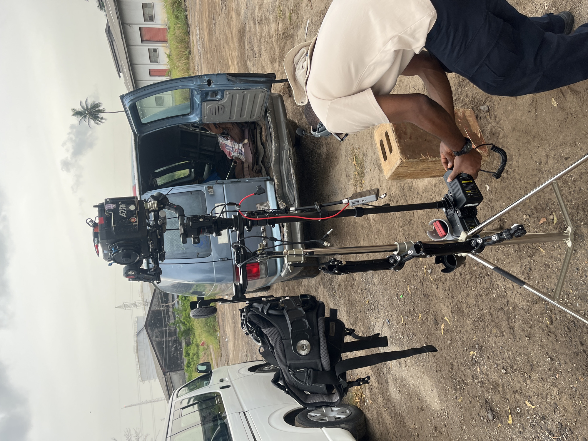 The image captures an outdoor film set environment in Lagos, Nigeria. A camera is mounted on a tripod next to two vans, one blue and one white. The door to the blue van is open. A person wearing light-colored clothing is working on the camera equipment. A wooden apple box is also present. In the background, there are buildings, including a white building with red doors and a palm tree. The ground is a mix of dirt and gravel. Overhead electrical wires are visible, suggesting a location within an urban or industrial area. The weather appears overcast.