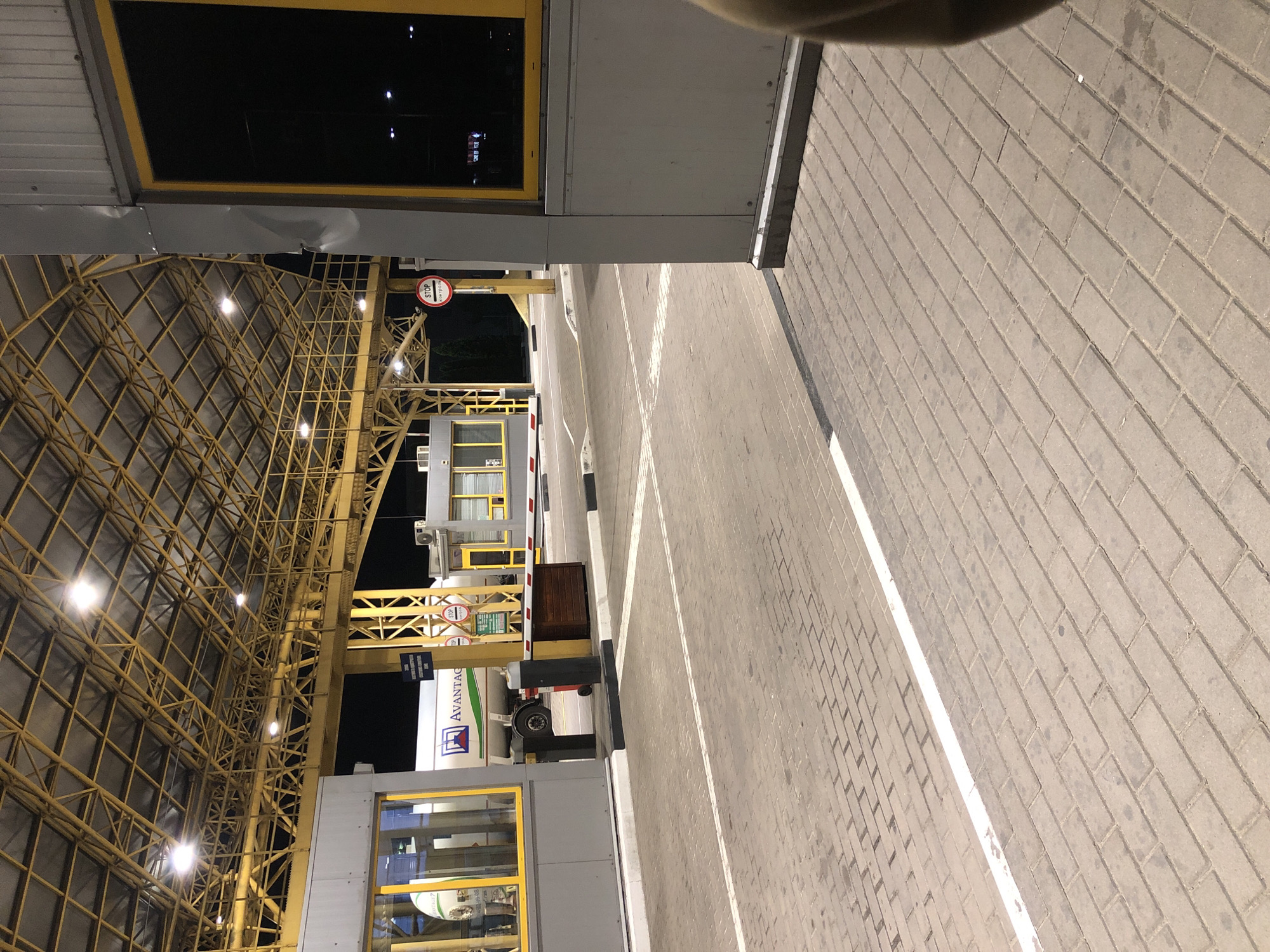 The image shows a border crossing checkpoint at night, likely at the Starovoitove, Ukraine location given the context. The scene is brightly lit by overhead fluorescent lights under a large, yellow metal canopy. In the foreground, there is a paved area made of interlocking bricks with white lines marking lanes. A raised white curb runs along the edge of the paved area.
To the left, there is a grey booth with yellow-framed windows. A truck with a white tanker trailer bearing the "AVANTAGR" logo is visible through the window of another booth further back. A red and white striped barrier arm is lowered across a lane.
On the right side of the image, a grey structure with a yellow-framed window is partially visible in the foreground, with a person's shoulder and arm appearing on the far right edge of the frame. A red circular sign with "STOP" and Cyrillic text is mounted on a yellow pole. Several other smaller buildings and structures are visible in the background, including what appears to be a control booth. The overall environment suggests a functional, active border crossing.
