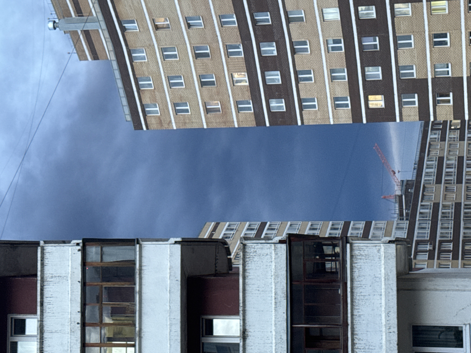 The image displays an upward-looking view of multiple multi-story buildings against an overcast, blue-grey sky. The photograph is rotated 90 degrees counter-clockwise, causing the tall structures to appear horizontally across the frame.
The upper portion of the image features a large building with a facade composed of light brown bricks or panels segmented by darker vertical stripes. Numerous rectangular windows with white frames punctuate the facade; some reflect the sky, while others show internal lighting or darkness. At the far left end of this structure, a small piece of rooftop equipment, possibly an antenna or vent, is visible. Thin, dark wires or cables stretch diagonally across the top-left against the sky.
In the lower part of the frame, a different building appears closer to the viewer. This structure has a lighter, possibly concrete or painted white, facade with large, dark-framed windows. The condition of these windows varies, with some reflecting light and others appearing worn or dark.
Nestled in the middle-right, partially obscured by the foreground buildings, another tall building with a similar aesthetic to the upper structure is visible in the distance. Behind it, a red construction crane extends towards the sky, indicating ongoing development or construction activities.
The sky occupies a significant central portion of the image, displaying a uniform deep blue-grey hue, characteristic of an overcast day. No people are visible. The scene captures an urban landscape in Perm, Russia, highlighting the utilitarian architecture common to the region and the presence of construction in the city.
