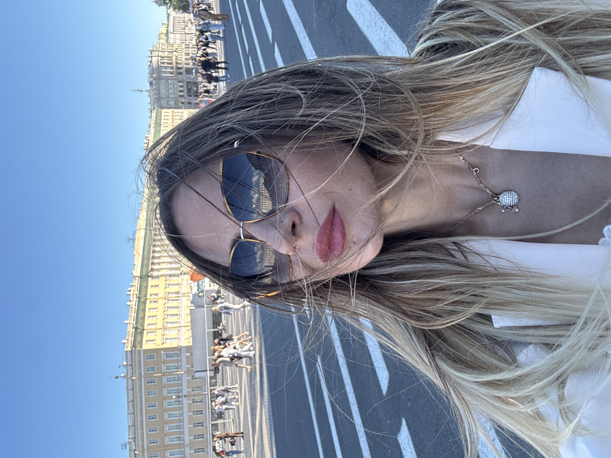 A woman is taking a selfie outdoors in front of a large, pale yellow building with a green roof and a colonnade. The building appears to be classical in architecture and is situated in a wide, open square. The sky is clear and blue, suggesting it is a sunny day. The woman is wearing oversized sunglasses with a gold frame, and her blonde hair is being blown by the wind. She has on a white blazer and a delicate necklace with a pendant. In the background, a crowd of people can be seen walking across the square, and there are white lines painted on the asphalt, possibly indicating a pedestrian crossing. The overall scene suggests a tourist location in Saint Petersburg, Russia, specifically Palace Square, with the General Staff Building in the background.