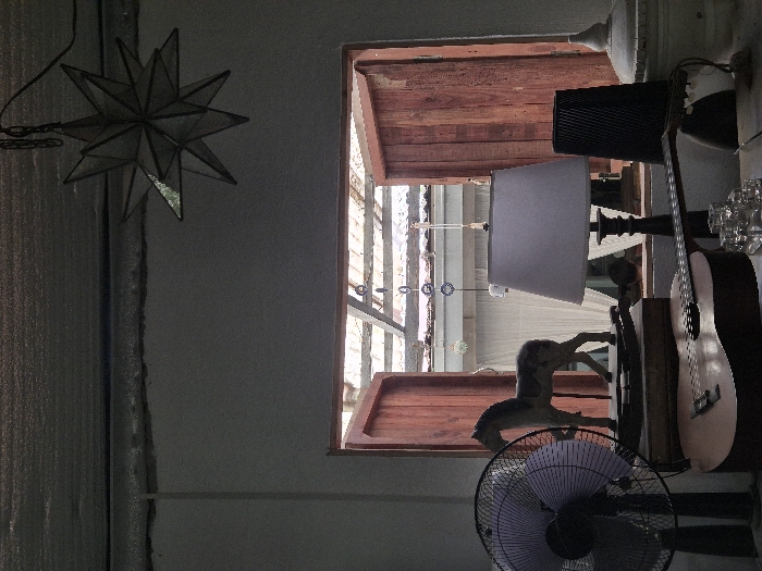 An interior scene captured in Silang, Philippines, presents a window looking out onto an outdoor or semi-outdoor area. The ceiling features a grey, possibly insulated surface with a linear structural element. A multi-faceted, star-shaped lantern, likely constructed from metal and glass, hangs from the ceiling by a chain and power cord on the upper right. Below, a light-colored wall forms the interior.
A wooden-framed window with two open, reddish-brown shutters dominates the mid-ground, providing a view of a brighter exterior. Through the window, corrugated roofing, possibly translucent, is visible along with hanging decorations including blue circular elements and white spherical objects. White sheer curtains are visible in the background, suggesting a covered outdoor living space.
In the immediate foreground, arranged on a surface in front of the window, are several objects. On the far left, a black standing electric fan with a light purple/grey protective grille is positioned vertically. To its right, a wooden rocking horse figurine rests on a dark wooden base. An acoustic guitar with a light-colored body and dark fretboard lies horizontally across the lower center. A table lamp with a white fabric shade and a dark, ornate base is situated to the right of the rocking horse. Behind the lamp, a dark, ribbed cylindrical object stands upright. On the far right, a white, finial-topped decorative urn or container is partially visible. A cluster of clear glass shot glasses or small cups is present on the surface in the lower right foreground. The scene is illuminated by natural light entering primarily from the window, creating a silhouette effect for some interior objects.