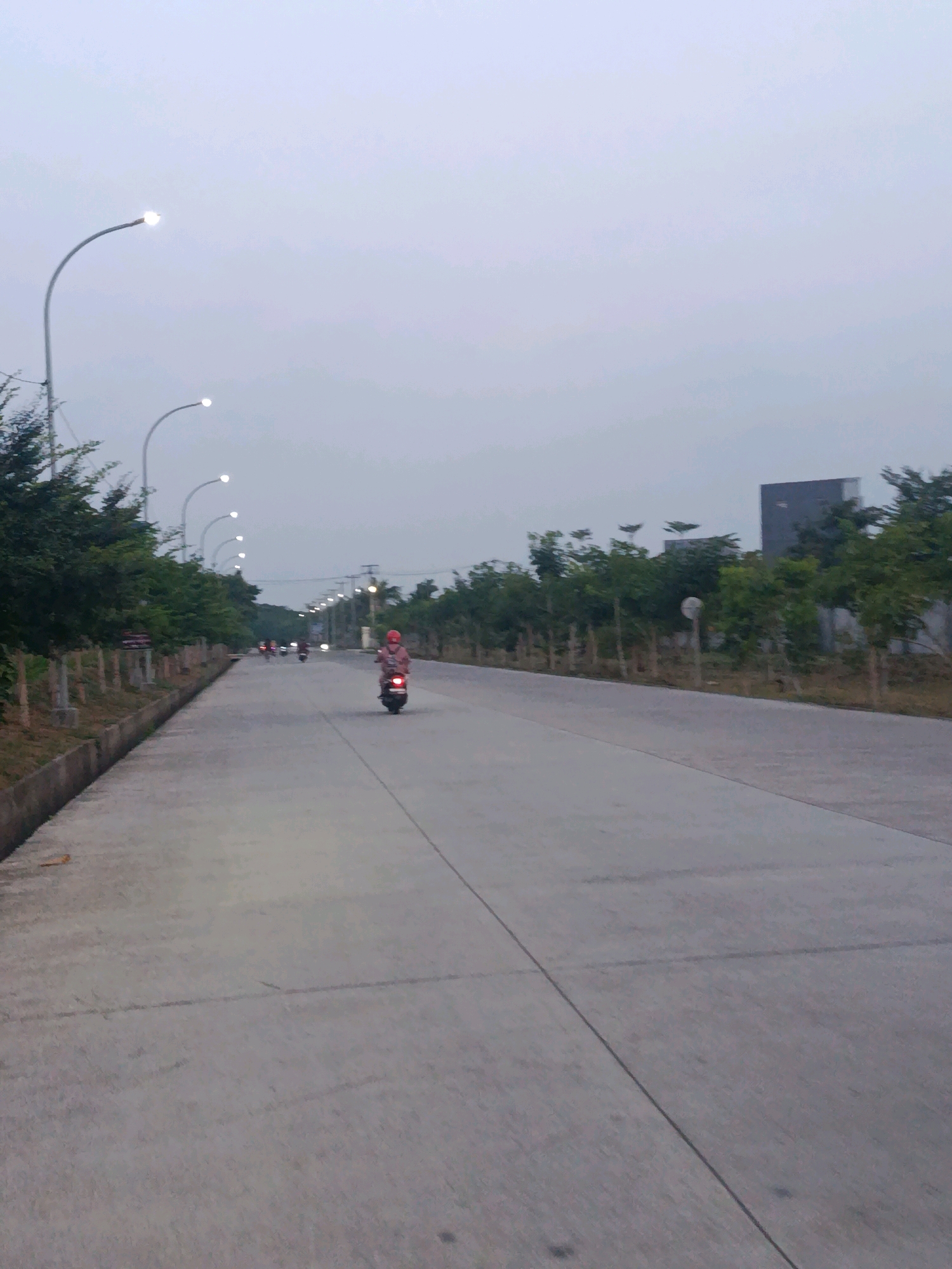 A person is riding a scooter on a paved road in Bunibakti, Indonesia, during dusk. The road is flanked by trees and streetlights. The sky is overcast.