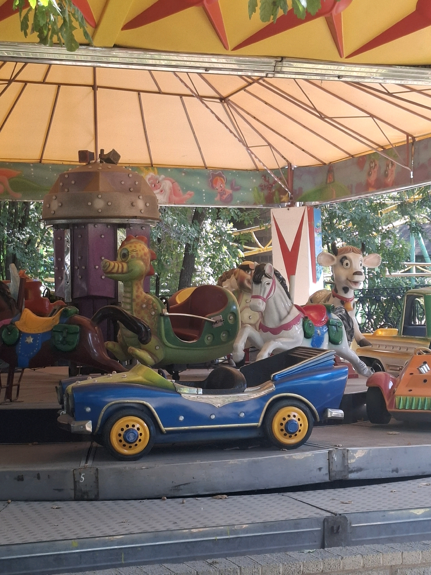This image is of a merry-go-round ride, likely at an amusement park or a fair, located in the city of Almaty, Kazakhstan. The merry-go-round features several colorful amusement ride vehicles, including a blue car with yellow wheels, a white horse with pink accents, a green dinosaur-like creature, and a brown horse. In the background, a cow figure is visible. The ride is covered by an orange and yellow awning, and lush green trees surround the area, suggesting an outdoor setting. The ride appears to be empty of any people.