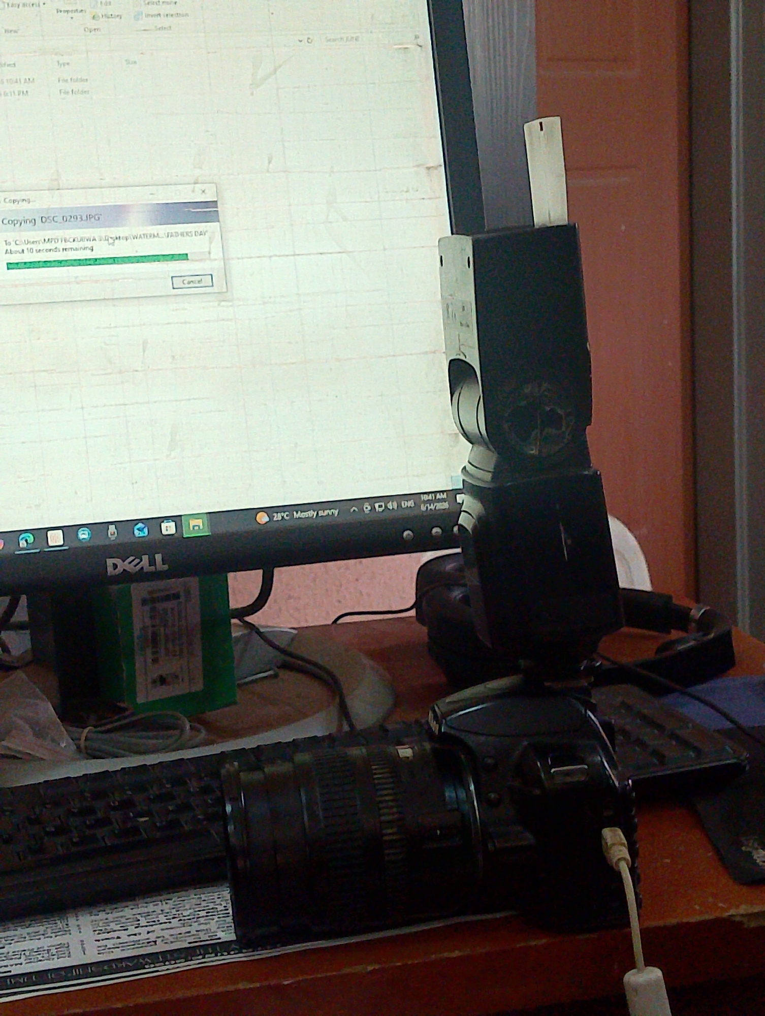 The image captures a close-up view of a desktop computer setup in what appears to be a residential or office environment in Kubwa, Nigeria. A Dell monitor displays a "Copying DSC_0293.JPG" progress window, indicating a file transfer is in progress. Below the monitor, a black keyboard is visible, and to its right sits a digital camera, likely a DSLR, with a lens attached. A flash unit is mounted on the camera's hot shoe, and a white cable is plugged into the camera. The background features a reddish-brown wall and part of a door. The scene suggests a digital photography workflow, possibly related to personal or professional use, taking place in an indoor setting in Kubwa.