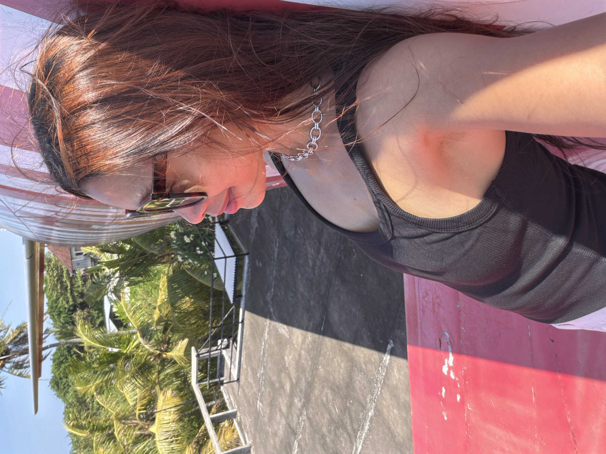 This is a close-up, side-profile photograph of a young woman with long, dark auburn hair illuminated by sunlight. She is wearing sunglasses with tortoise-shell frames and a silver chain necklace. Her head is tilted, and she appears to be resting on something pink and textured, possibly a lounge chair or the side of a structure.
The background suggests a tropical setting with lush green palm trees and foliage visible to her left. A gray, weathered surface, possibly pavement or a roof, occupies the right side of the image, with a stark shadow cast across it, indicating a sunny day. A metal ladder or staircase is also visible in the background, leading up towards the foliage and what appears to be a building.
The overall mood is relaxed and casual, with the bright sunlight and tropical greenery suggesting a warm, outdoor environment. The focus is on the woman and her immediate surroundings, with the backdrop providing context of a potentially resort-like or coastal location in Blahbatuh, Indonesia. The image is taken from a low angle, looking up at the woman. There is no visible text in the image.
