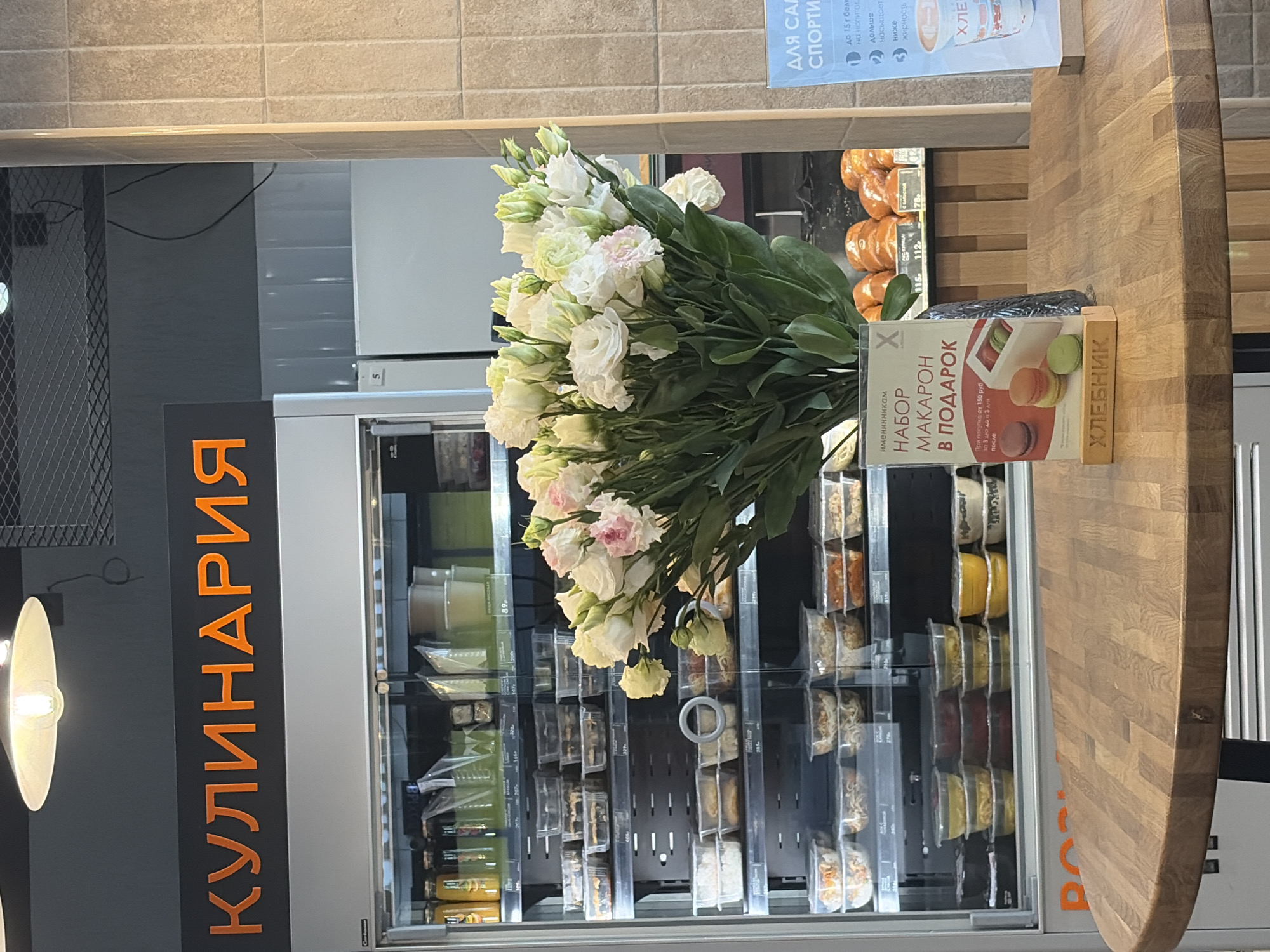 The image captures the interior of a culinary establishment, likely a shop or café in Saint Petersburg, Russia, evidenced by the Cyrillic script on the "КУЛИНАРИЯ" sign. The scene features a refrigerated display case stocked with prepared meals and snacks. A bouquet of white flowers is prominently positioned, partially obscuring the refrigerated case. Adjacent to the flowers, on a wooden counter, rests a marketing display advertising "МАКАРОН" (macarons) in a gift set, with the text "ХЛЕБНИК" (bread maker) beneath. The upper section of the image shows a promotional banner with details for "ДЛЯ САЛ СПОРТИ" (for sport salads) indicating health-conscious food options. The lighting is bright, suggesting an artificial source. The focus is on the culinary offerings and the flowers, possibly arranged for decorative or promotional purposes.
