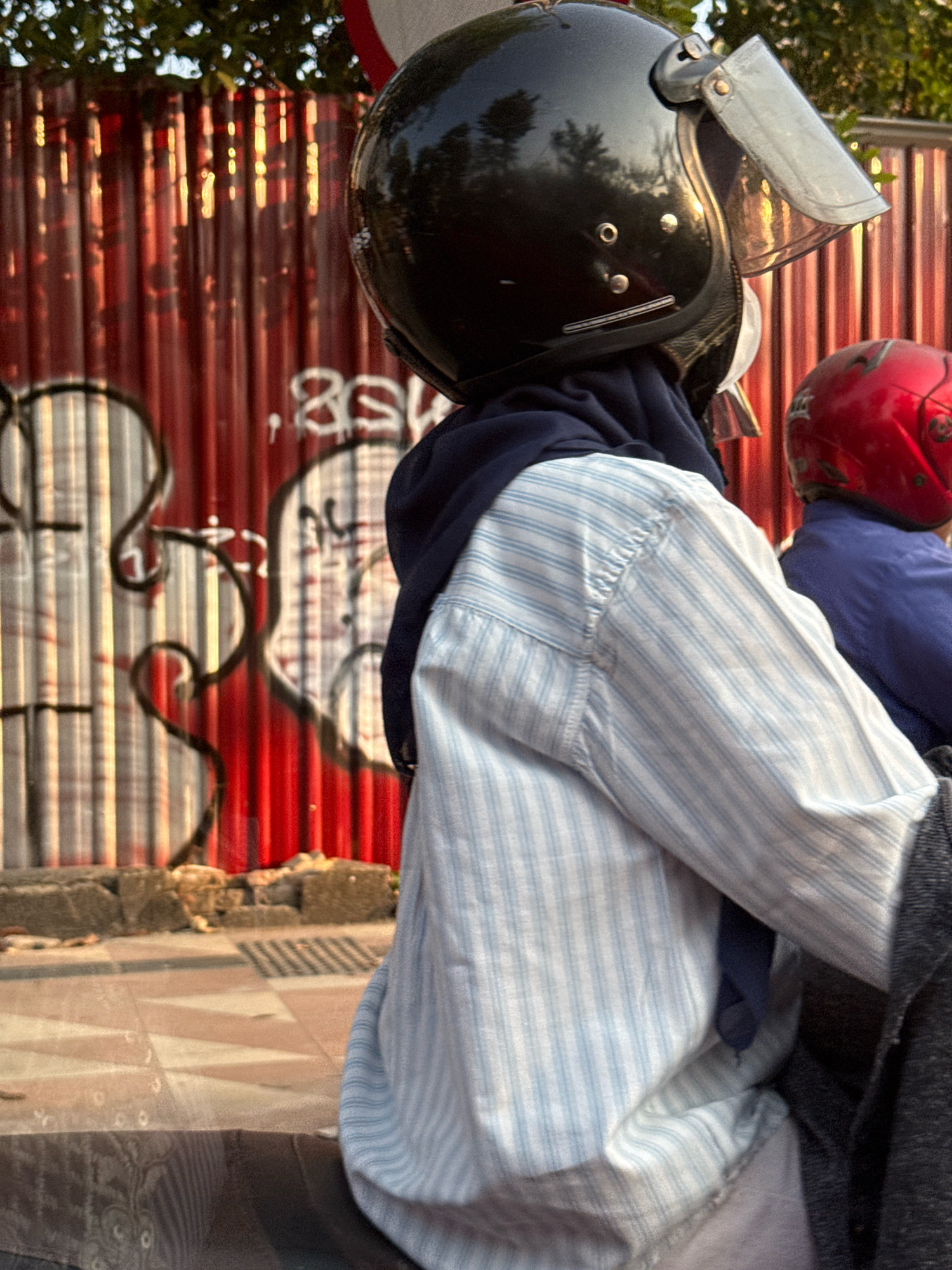 In Surabaya, Indonesia, this image captures a close-up view of two individuals on a motorcycle, seen from the side. The person in the foreground wears a black helmet with a clear visor and a navy blue head covering under a light blue and white striped long-sleeved shirt. Their right arm is visible, and they appear to be holding onto the motorcycle. Behind them, a second person wears a red helmet and a dark blue jacket. They are positioned in front of a corrugated red metal fence covered in graffiti. The ground is paved with a mosaic-like pattern, and sunlight casts a warm glow on the scene, highlighting the textures of the fence and the clothing.