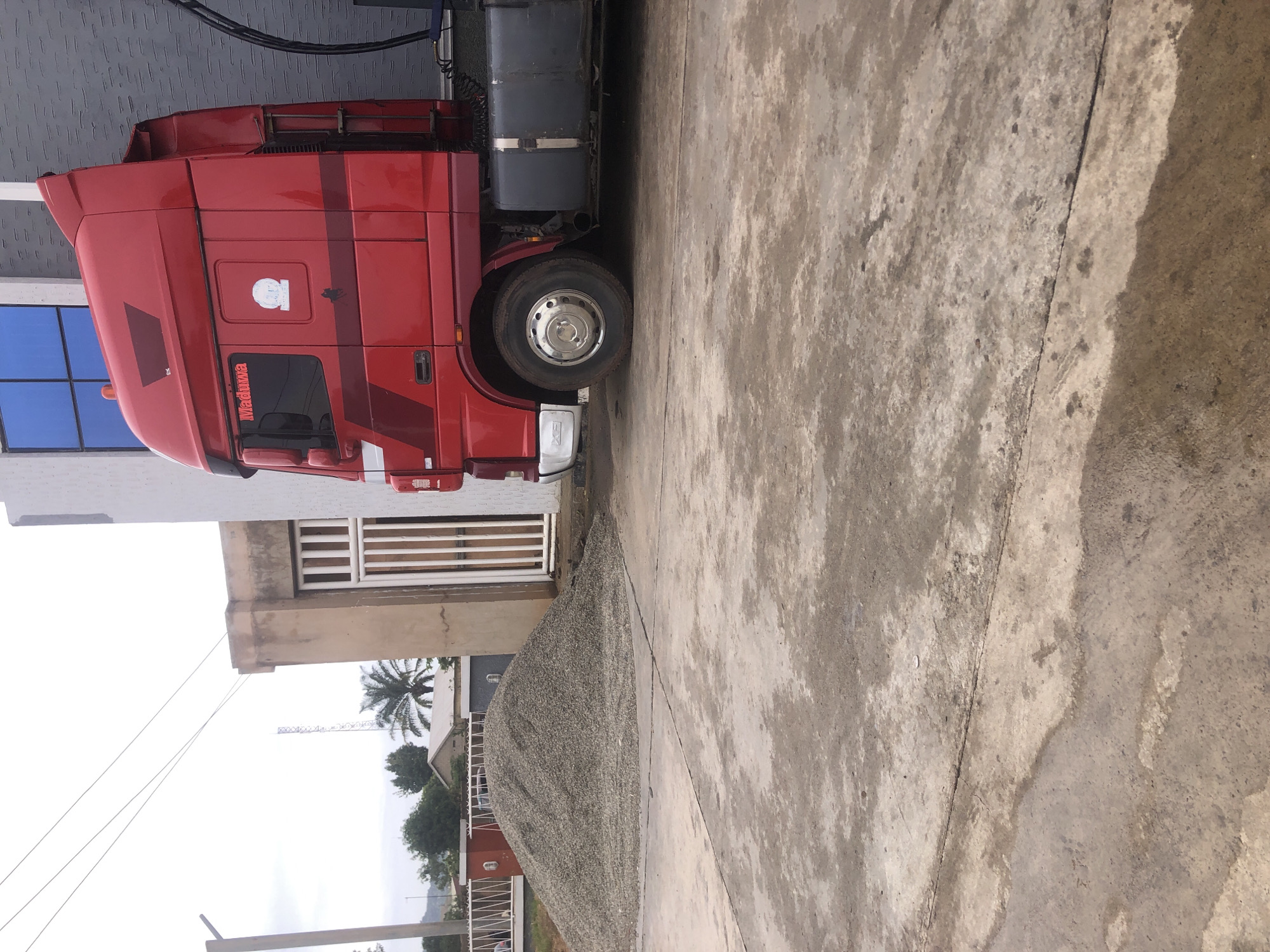 In New Stadium, Nigeria, a bright red truck is parked beside a building with a white and tan facade. The truck's cab features the name "Maduwa" in red lettering and a circular emblem. A large pile of gravel sits in front of the building, near a white gate. The ground is a concrete surface that appears wet and stained in patches. In the background, trees and a cell tower are visible against a cloudy sky. The overall scene suggests an industrial or construction-related location.