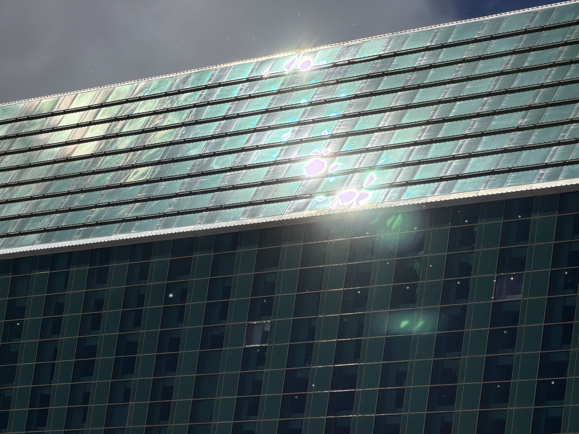 The image shows a section of a modern building in Moscow, Russia, with a distinctive roof and glass facade. The roof appears to be composed of numerous rectangular or square tiles, possibly solar panels, arranged in rows. Sunlight reflects strongly off the surface of the tiles, creating bright flares. Below the roof is a section of the building with a grid pattern of glass windows. One window is open. The sky is partially cloudy.