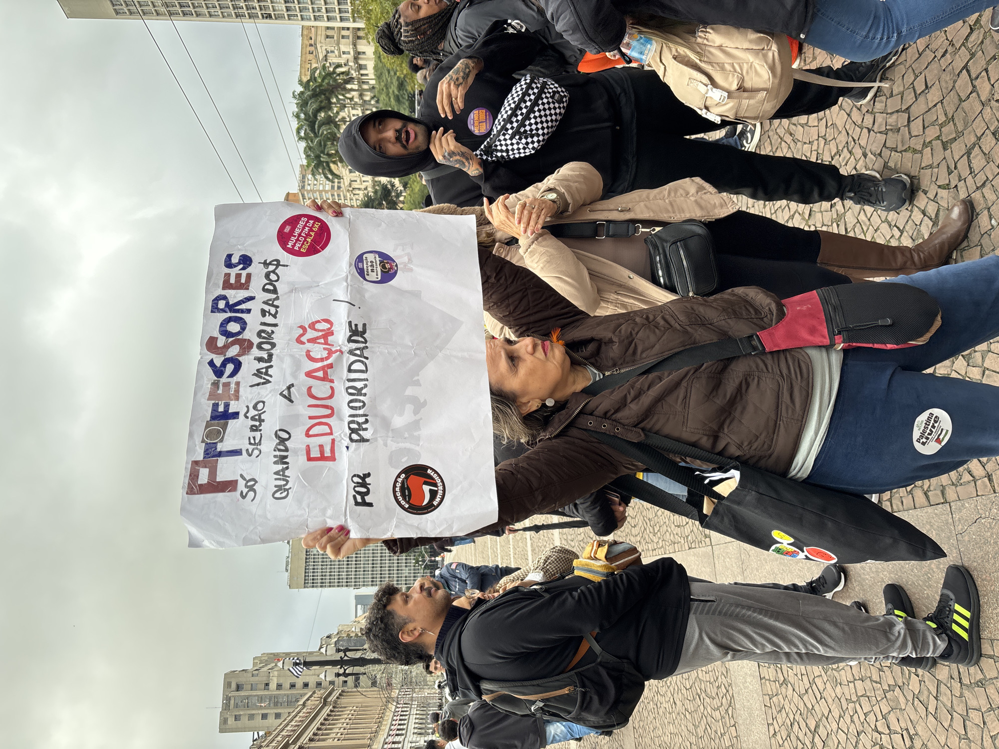 A medium shot shows a group of protestors holding a sign in Portuguese reading "PROFESSORES, SÓ SERÃO VALORIZADO$ QUANDO A EDUCAÇÃO FOR PRIORIDADE!" (TEACHERS WILL ONLY BE VALUED WHEN EDUCATION IS A PRIORITY!). The sign also includes stickers and political slogans.
The protestors are diverse in age and dress. Some are wearing casual clothing, while others have political stickers on their clothing and bags. The background features buildings and street elements, likely in a public square or street setting. The scene appears to be a political demonstration or protest. Based on the buildings and cobblestone ground, the location is likely in the city of São Paulo, Brazil.