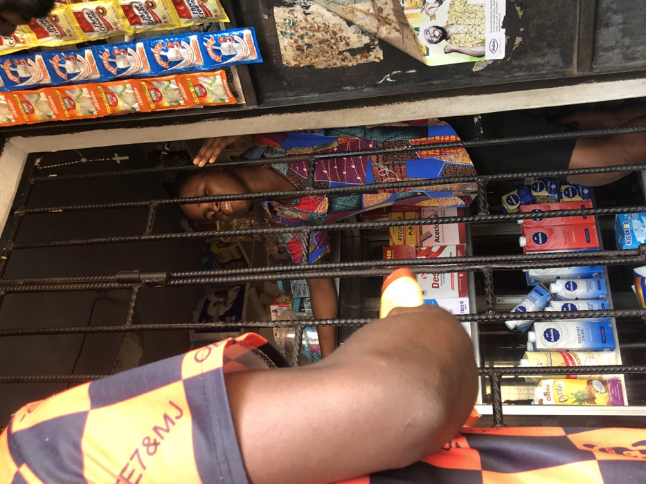 Through a metal-barred window, a woman in a colorful patterned shirt is visible behind shelves stocked with various items. These include beverage cartons like "Chivita" and "Kisski", dairy products labeled "Yogurt Drink", and boxes of "Acecloa" and "Rellx." On the outside of the window are products like "Three Crowns" milk powder. A leg in checkered orange and black shorts is positioned in the foreground, partially obscuring the view.